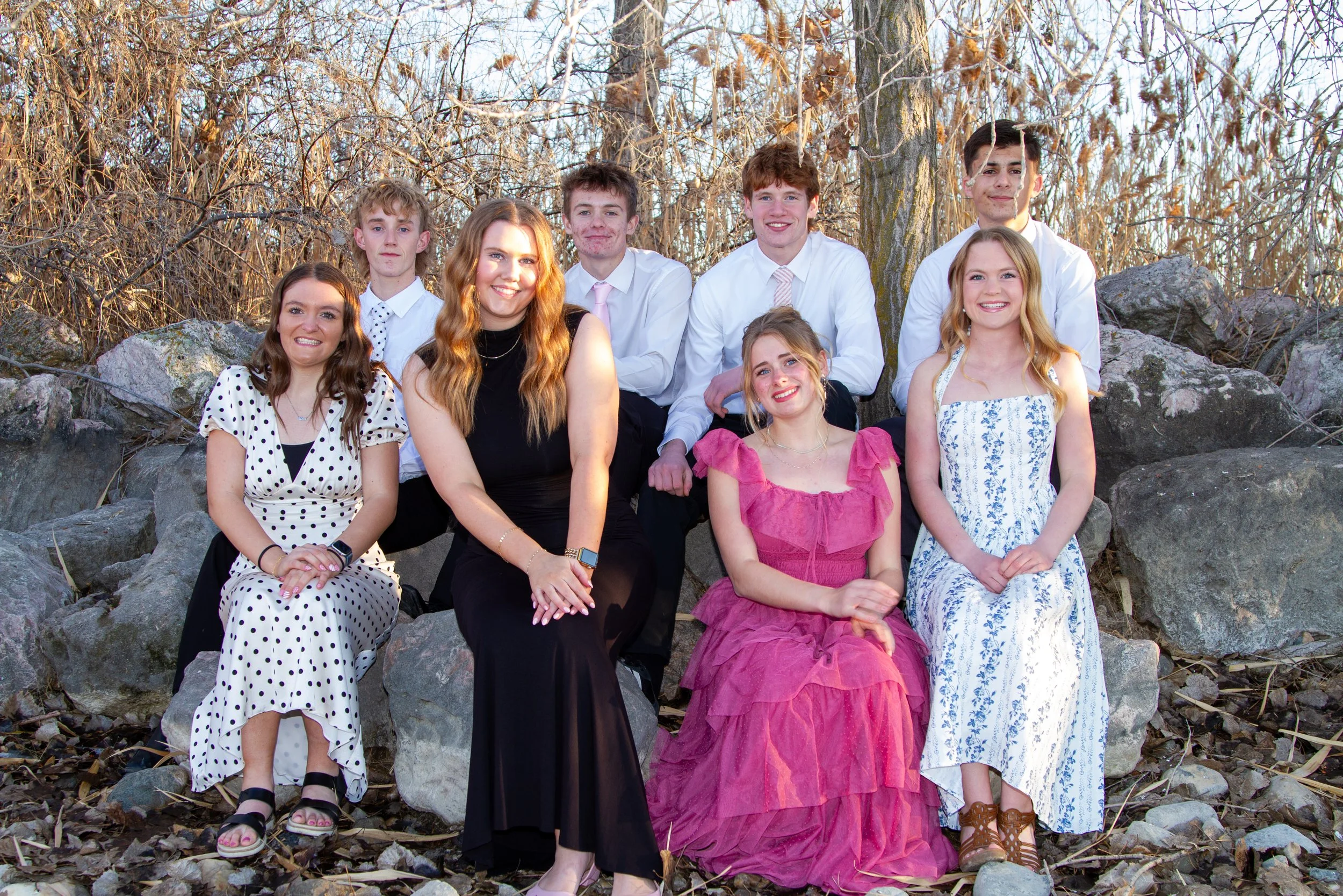A group of nine young people, five women and four men, posing outdoors on rocks with leafless trees in the background. They are dressed in semi-formal attire, with the women wearing dresses and the men in shirts and ties. The sunlight suggests it mig