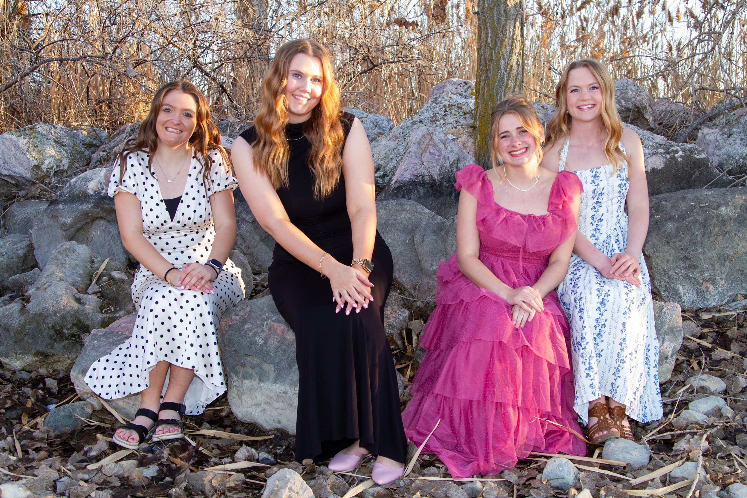 Four women sitting on rocks in a wooded outdoor area during daytime, dressed in summer dresses and smiling at the camera.