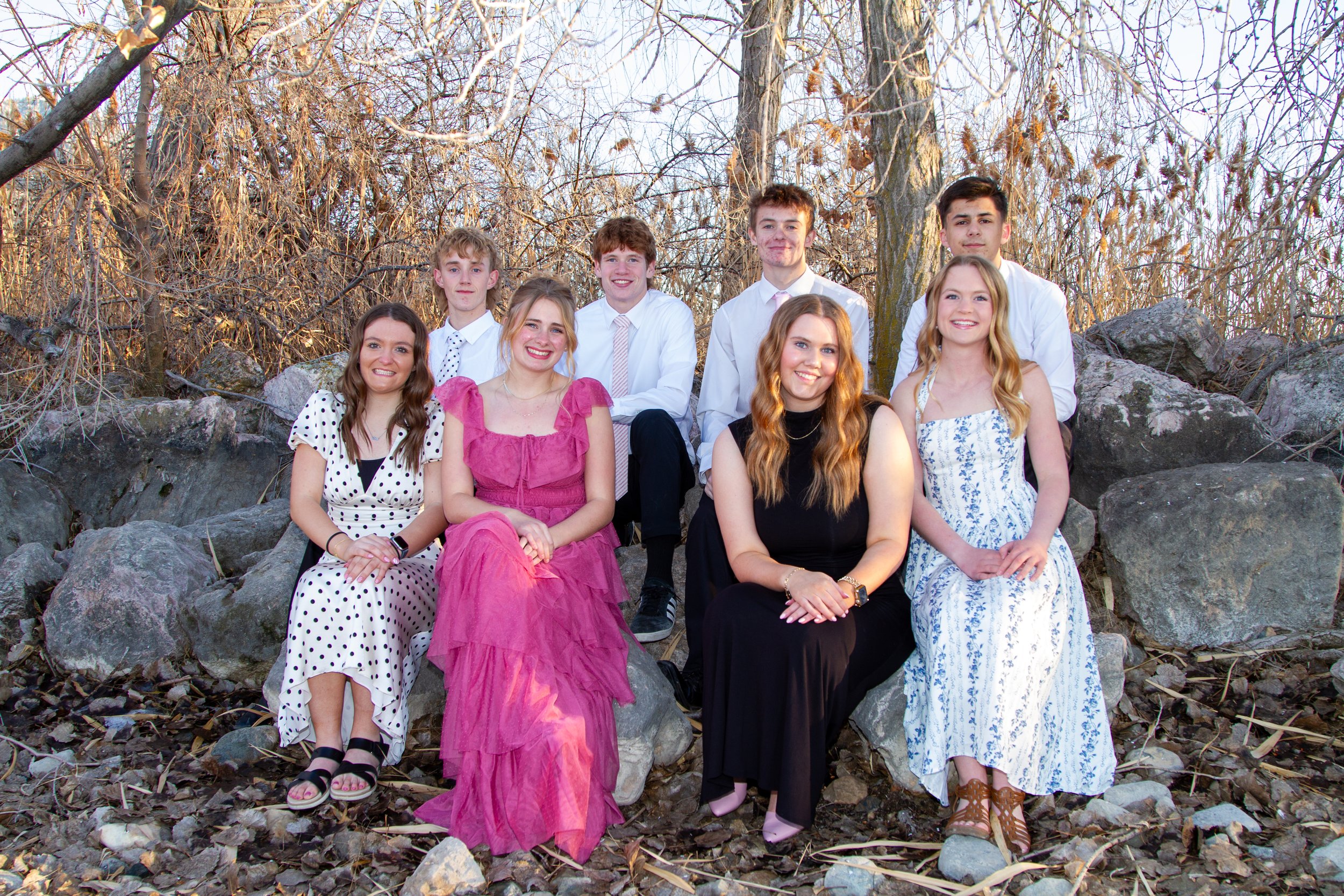 Group of nine teenagers sitting and standing on rocks in an outdoor setting with leafless trees in the background.