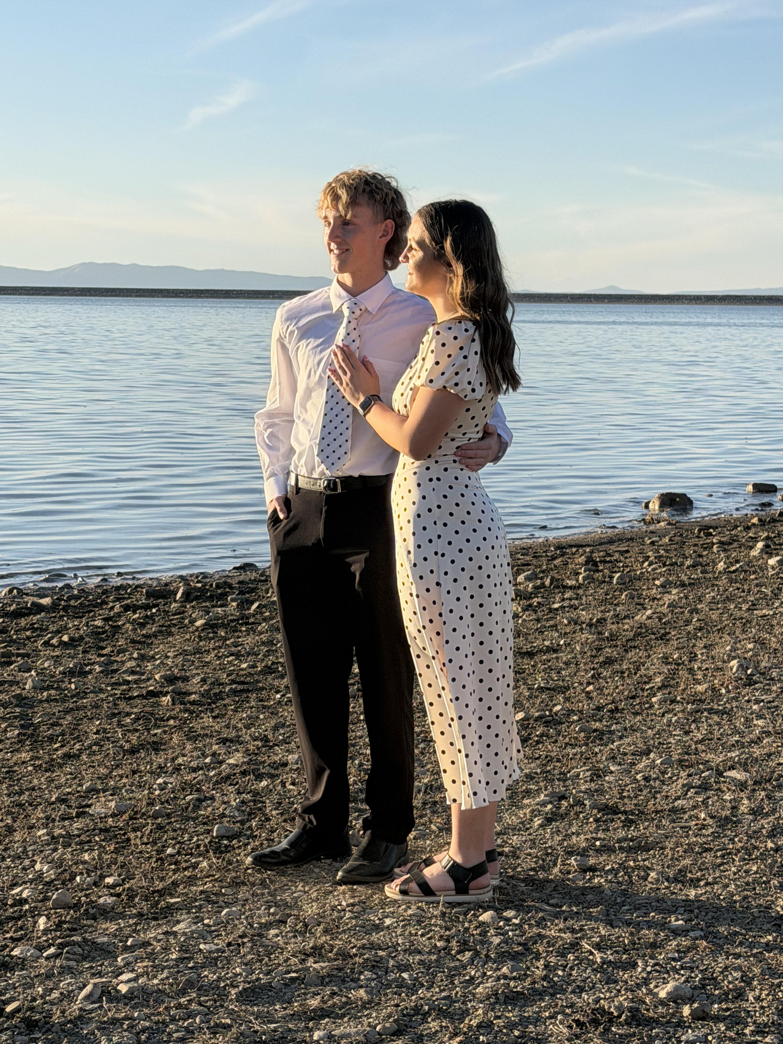 A young couple standing on a rocky shoreline near a body of water, dressed in semi-formal clothes, with the man in a white shirt and polka-dot tie, and the woman in a polka-dot dress, both gazing into the distance during sunset.