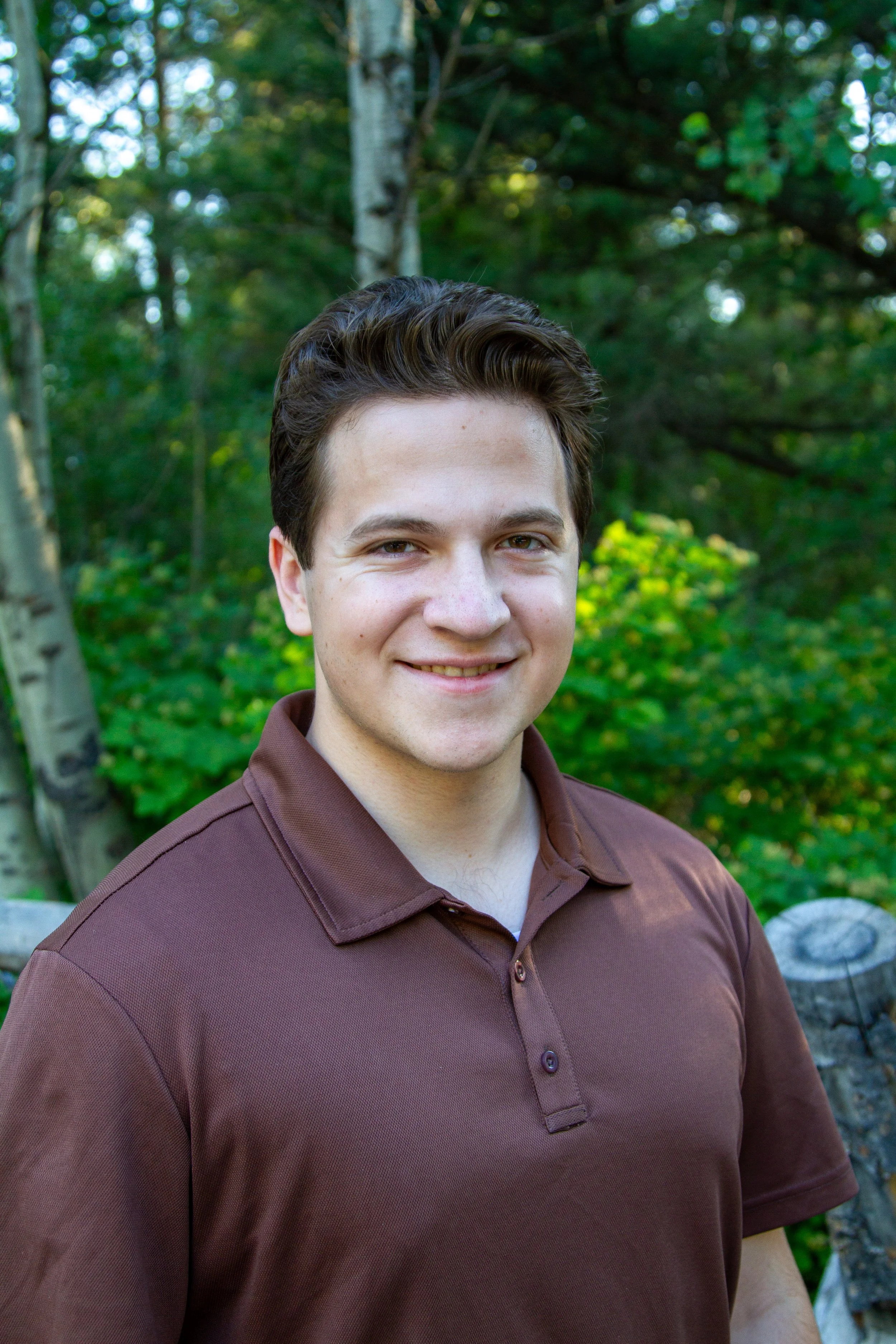 A young man with dark brown hair smiling outdoors, wearing a maroon polo shirt, with green trees and foliage in the background.