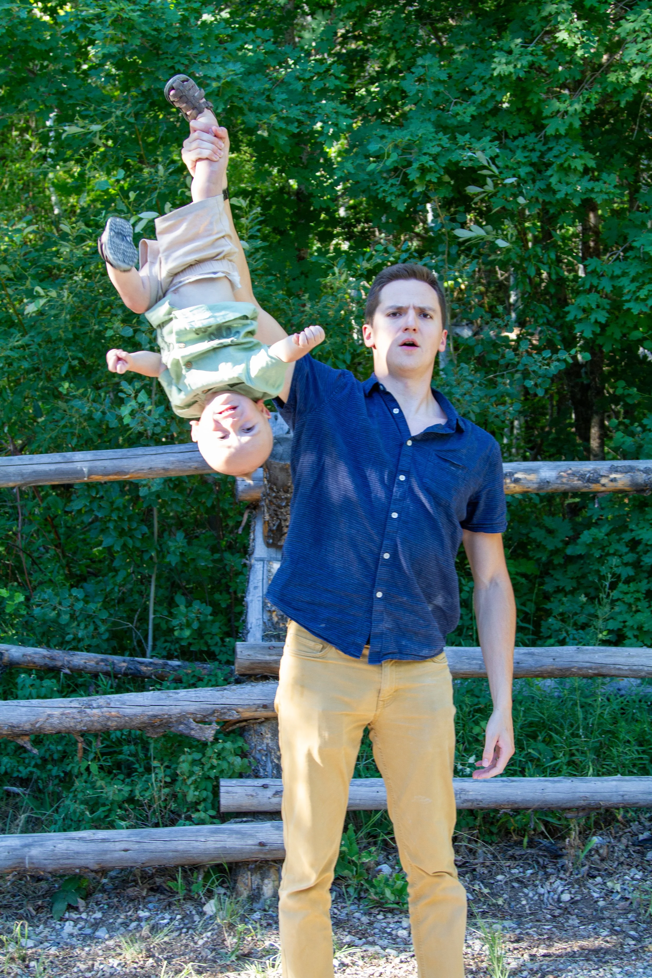 A man with short dark hair, wearing a navy blue button-up shirt and yellow pants, standing outdoors in front of a wooden fence with green trees in the background. He is holding a child upside down by the legs, with the child's head near the ground an
