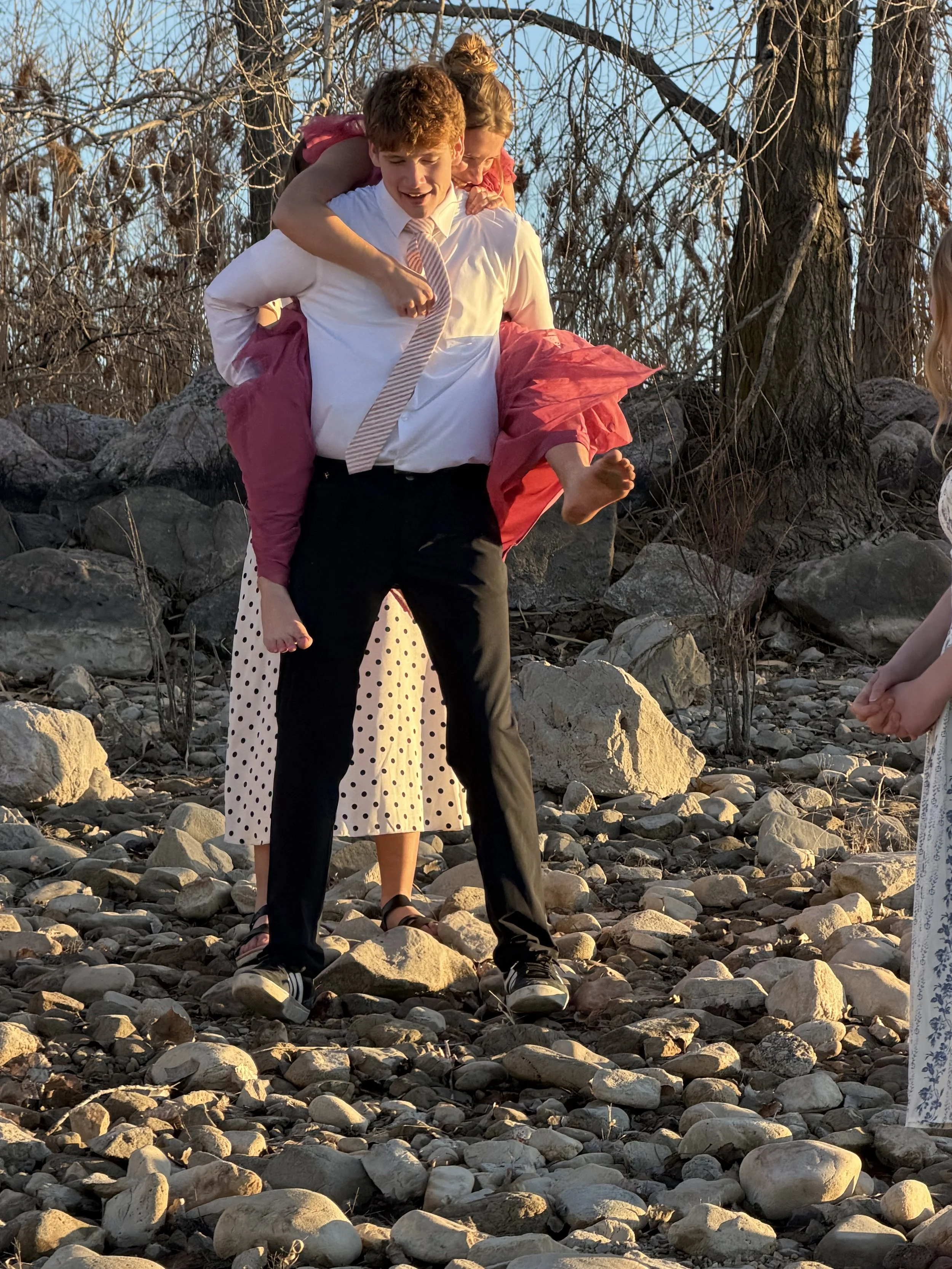 A man in formal attire carrying a young girl on his shoulders, outdoors on rocky terrain with trees in the background, during sunset.