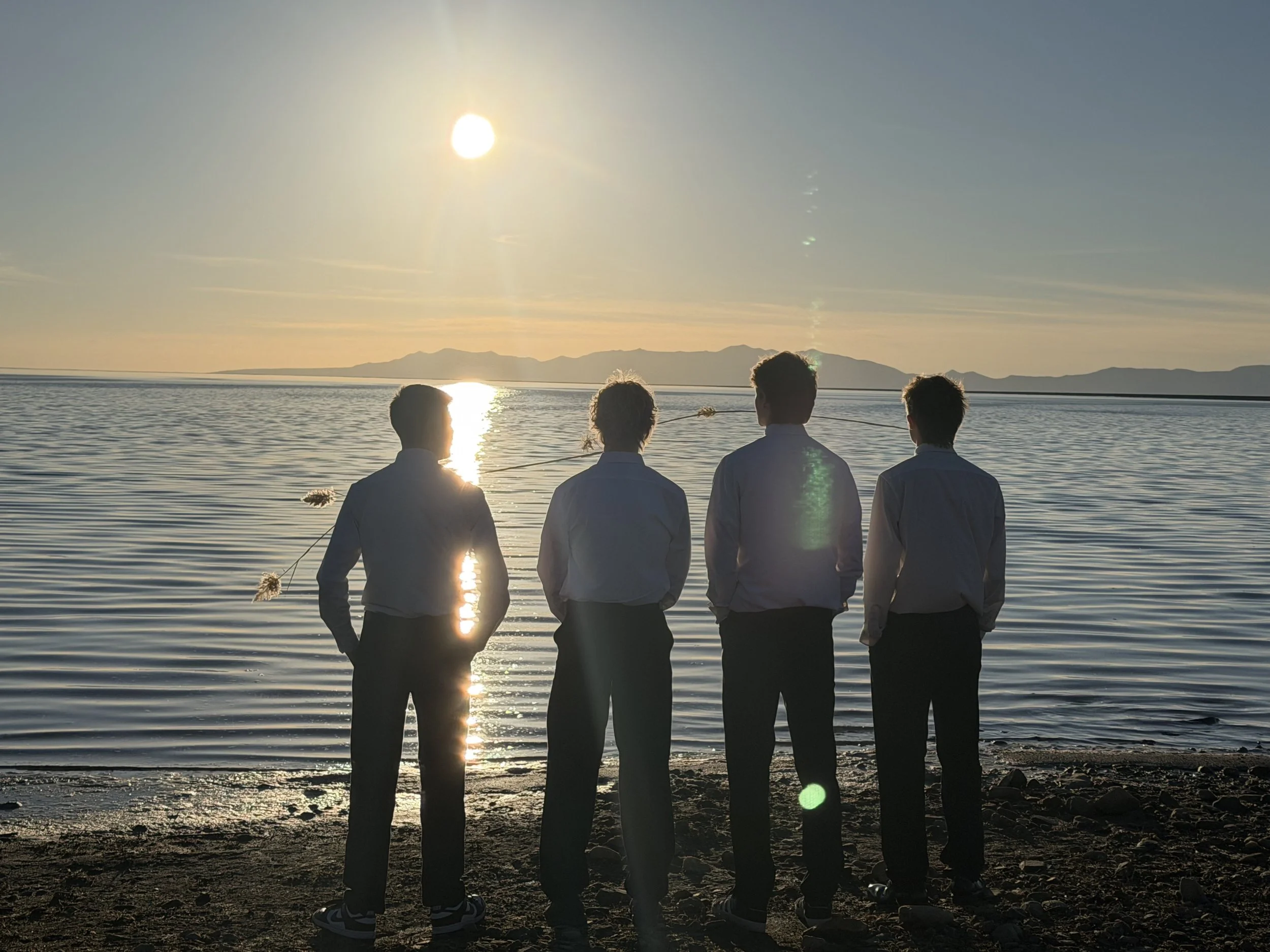 Four men in formal attire standing on a rocky beach at sunset, facing a calm body of water with mountains in the distance.