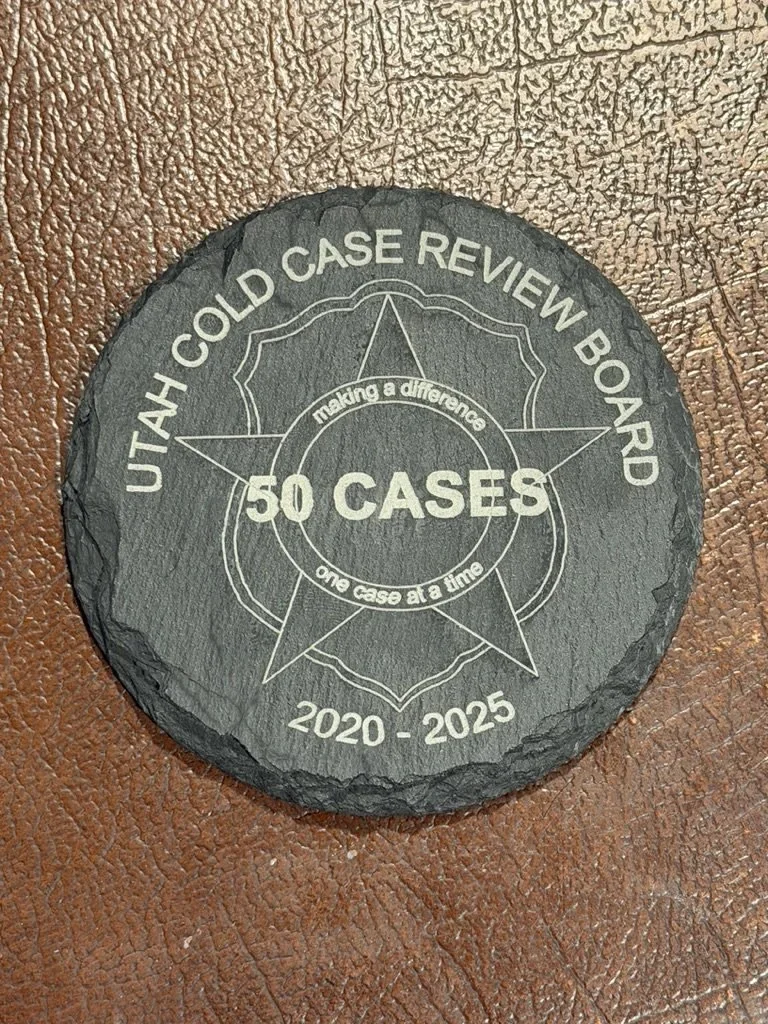 A round black stone with a logo for Utah Cold Case Review Board, featuring a star and the text "50 Cases, one case at a time, making a difference," with the years 2020-2025 etched on it, placed on a brown textured surface.