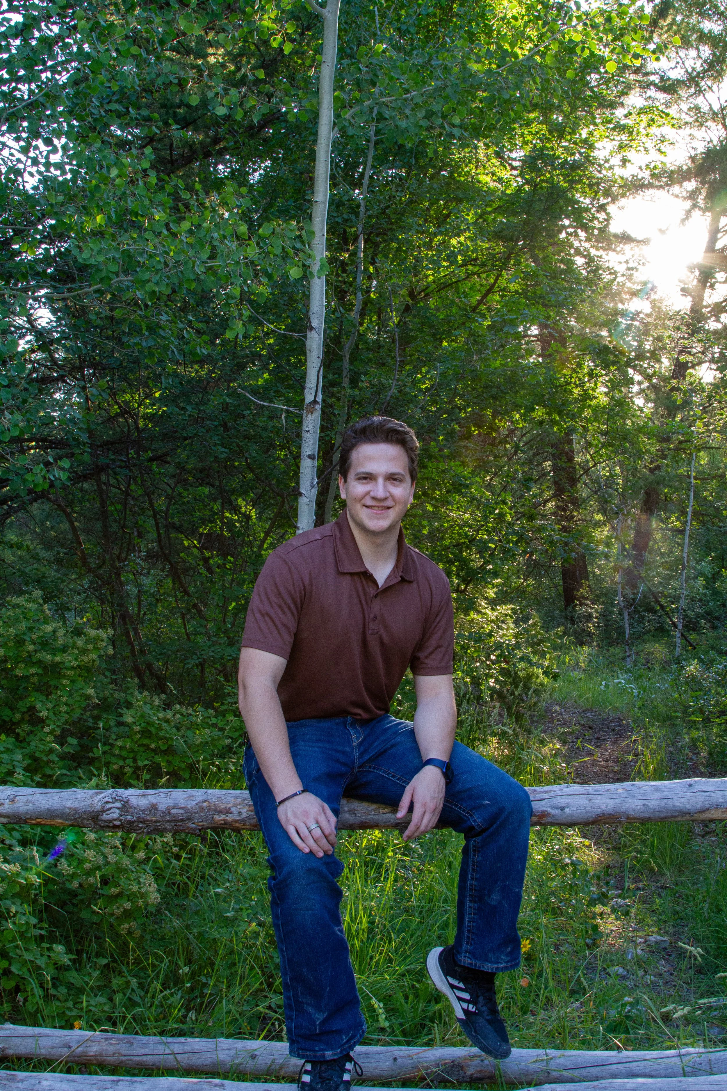A young man smiling and sitting on a log in a forest with green foliage and sunlight in the background.