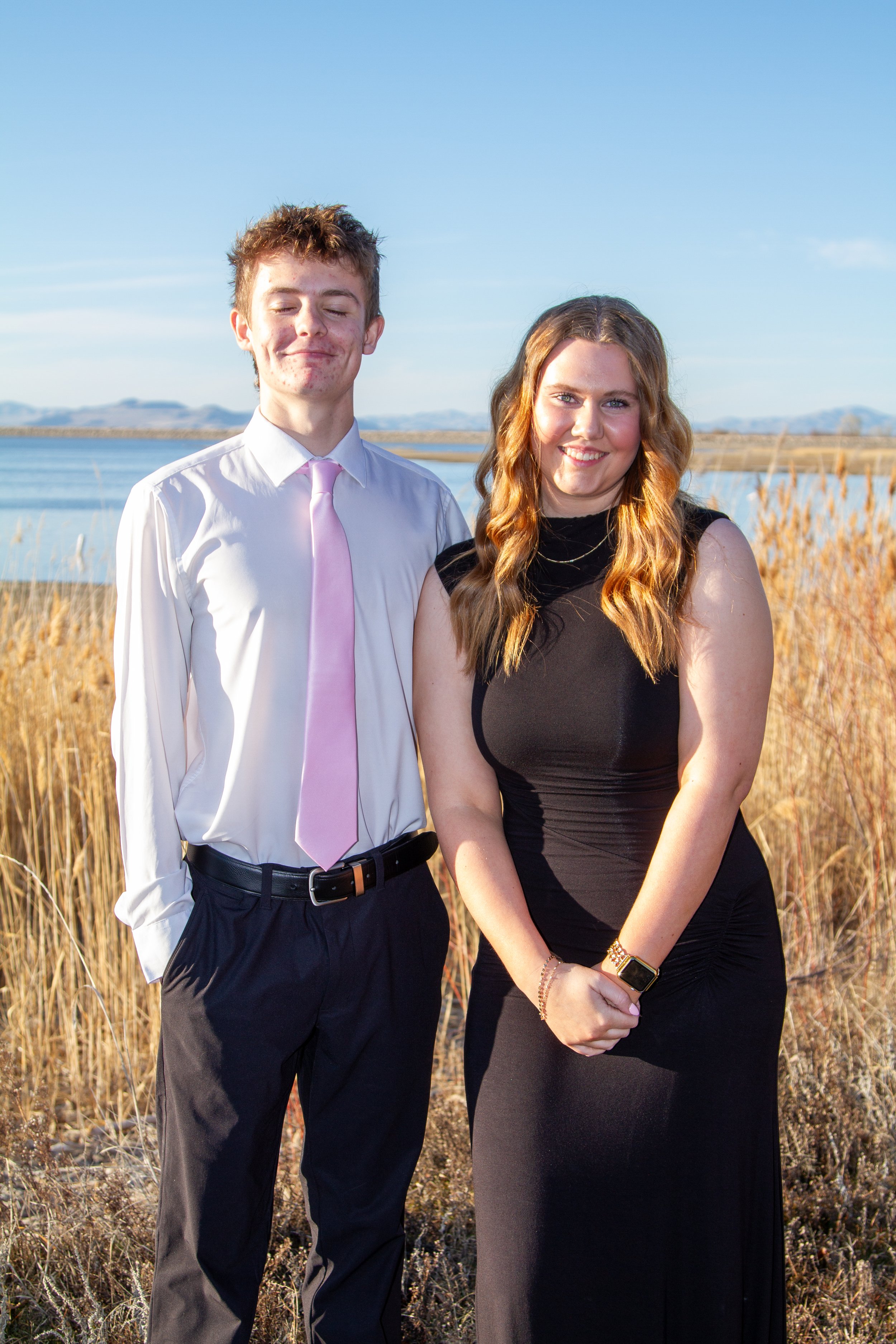 Young man and woman standing outdoors near a body of water, dressed formally for an event, with golden grass and a clear blue sky in the background.