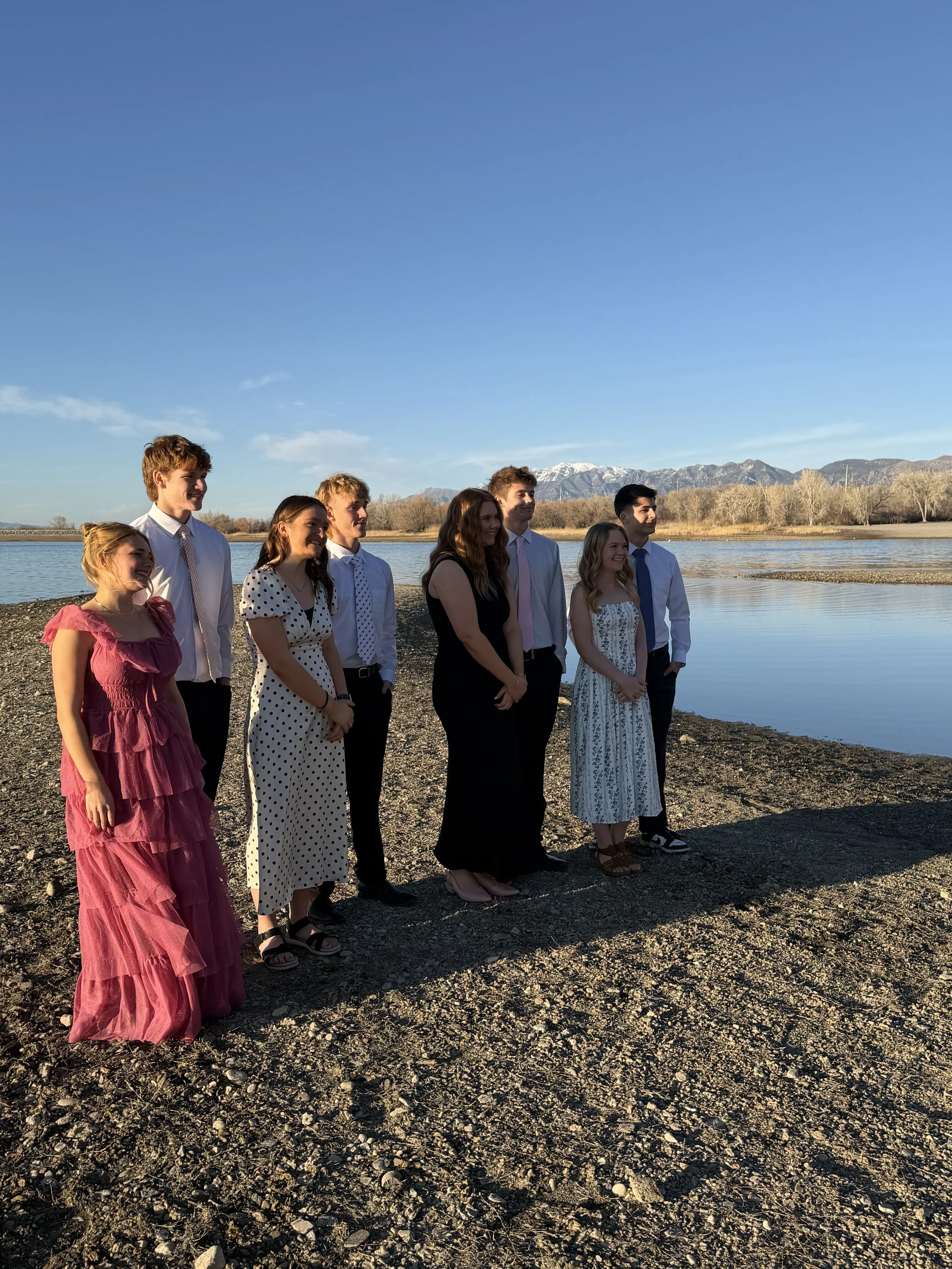 Group of nine young adults standing on rocky shore near water with mountains in background