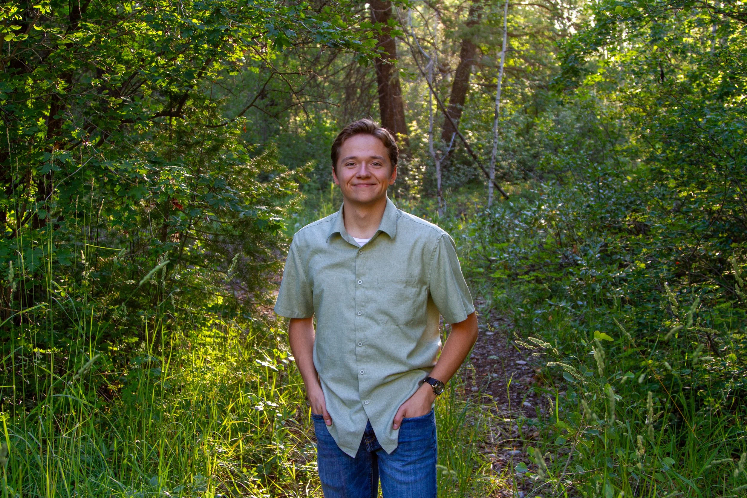 A young man smiling and walking in a wooded area with lush green foliage during daytime.
