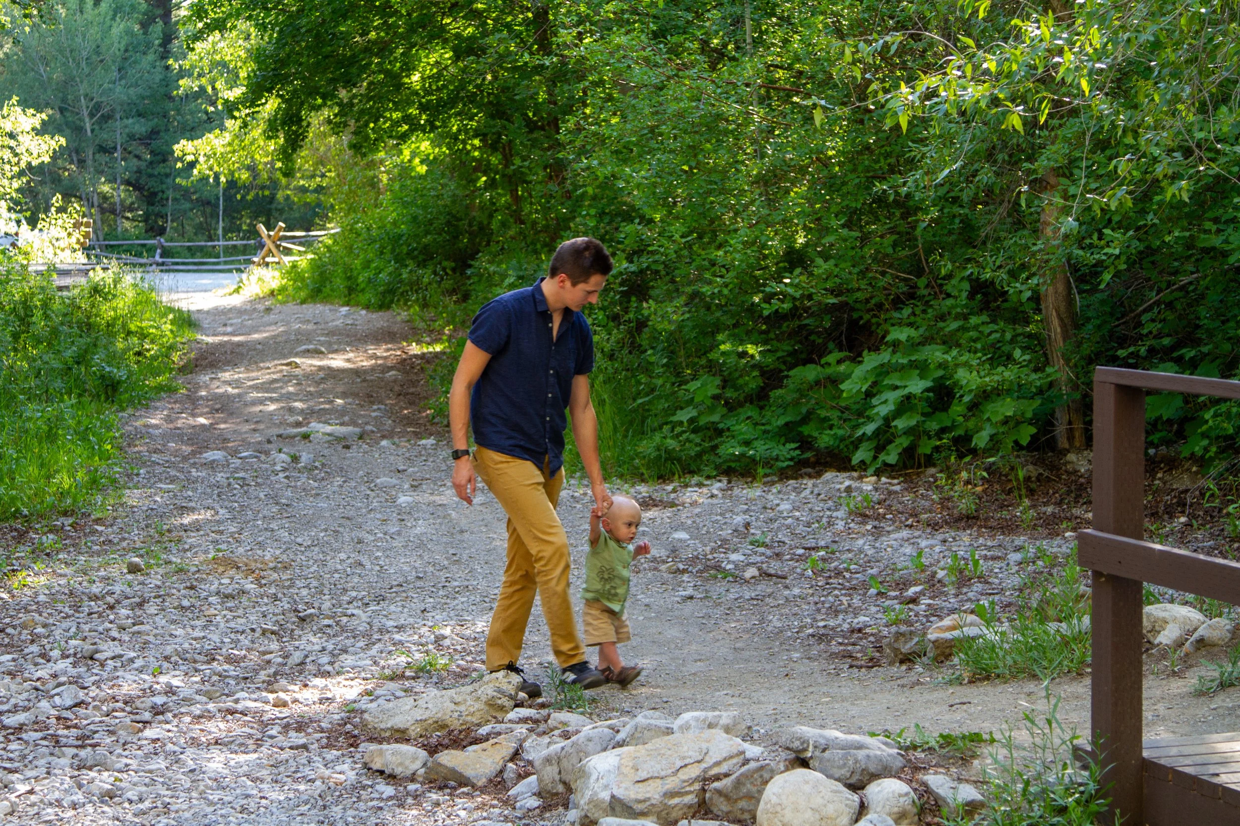 A man with short dark hair dressed in a blue shirt and khaki pants is walking with a young child on a rocky outdoor trail surrounded by lush green trees and foliage.