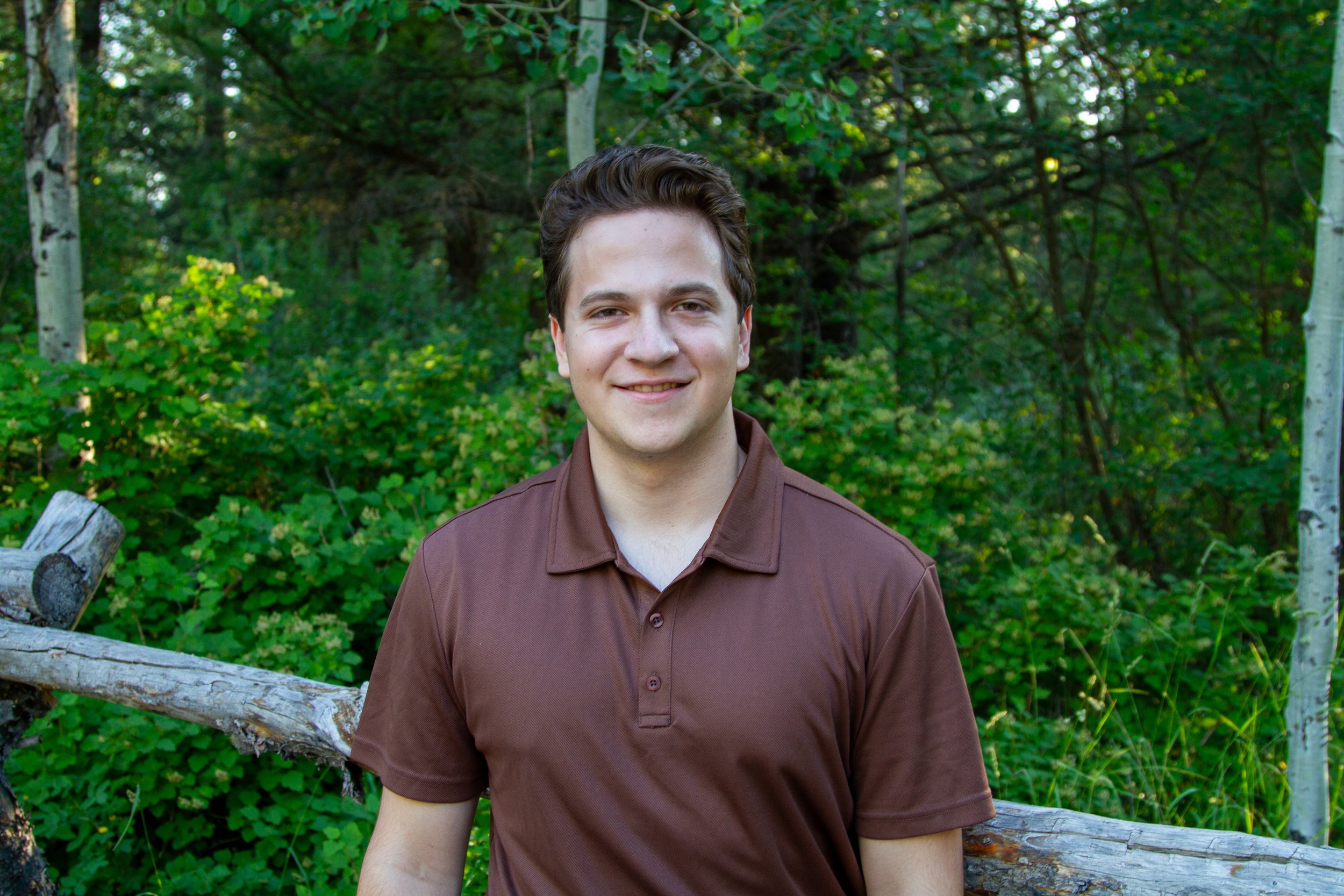 A young man with dark hair smiling outdoors in front of green trees, bushes, and a fallen tree trunk.