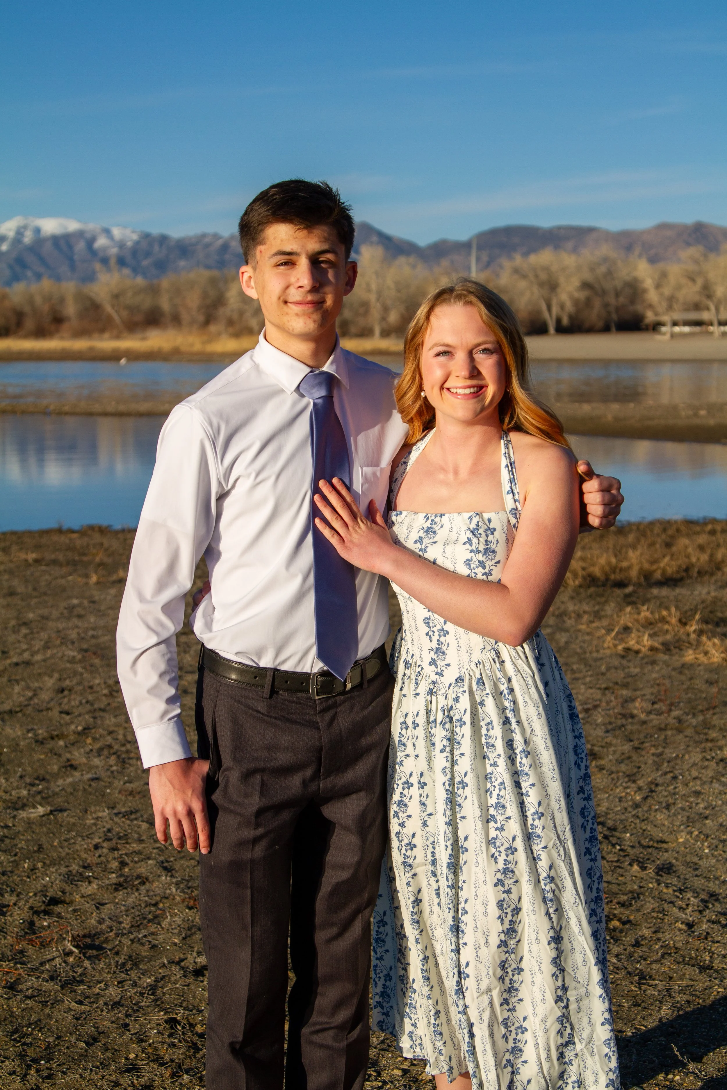 A young man and woman standing outdoors near a body of water, with mountains in the background. The man is wearing a white shirt and tie, while the woman is in a white and blue floral dress, both smiling at the camera.