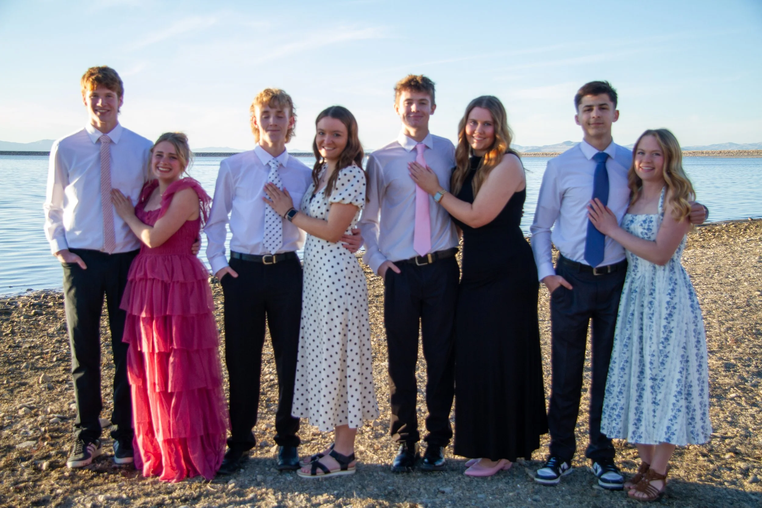 Group of eight teenagers standing on a rocky beach near a body of water, dressed in semi-formal attire, with clear skies and distant mountains in the background.