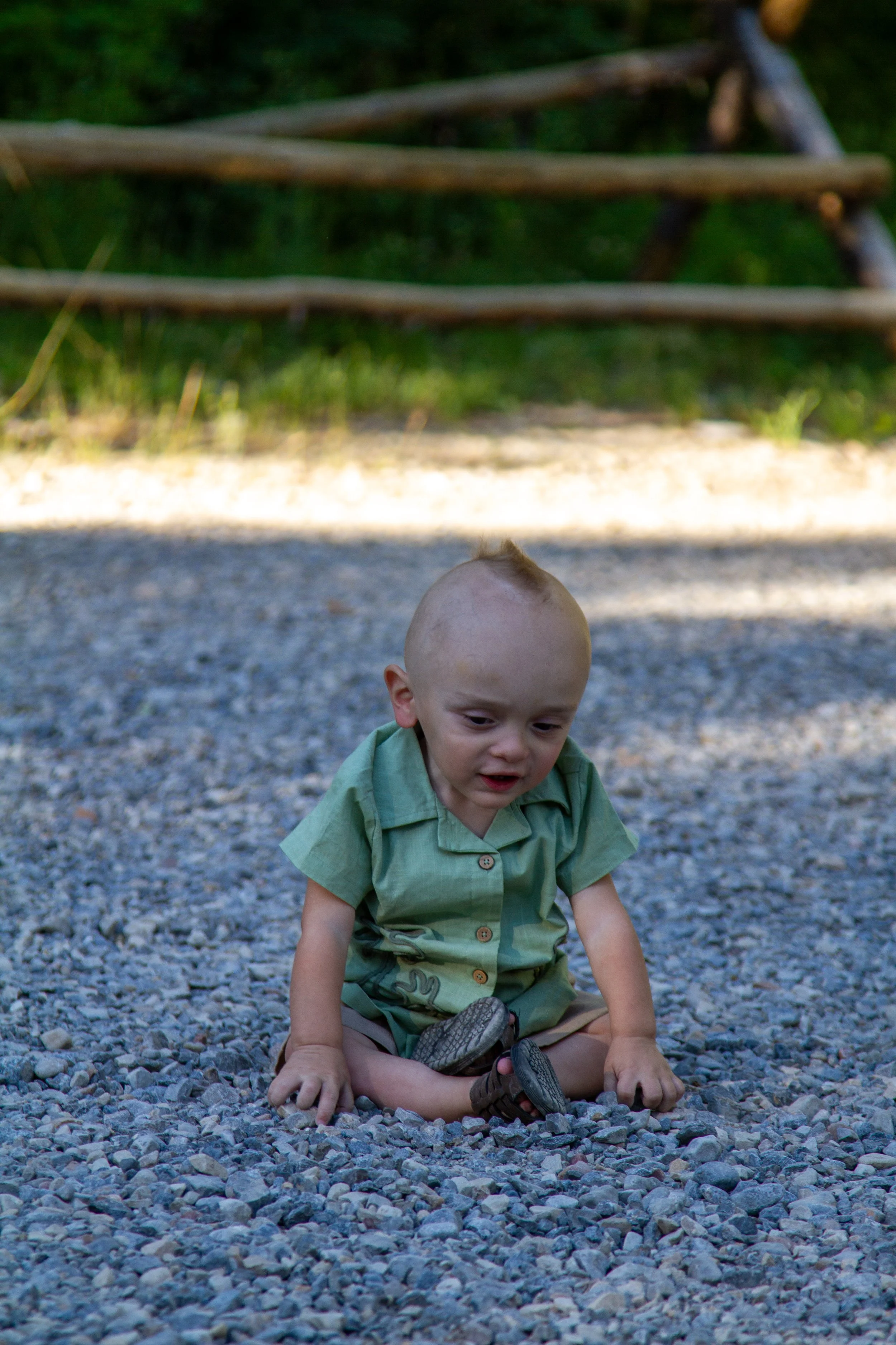 A young boy with a shaved head sitting on a gravel ground, holding a sandal and a shoe, with a wooden fence and greenery in the background.