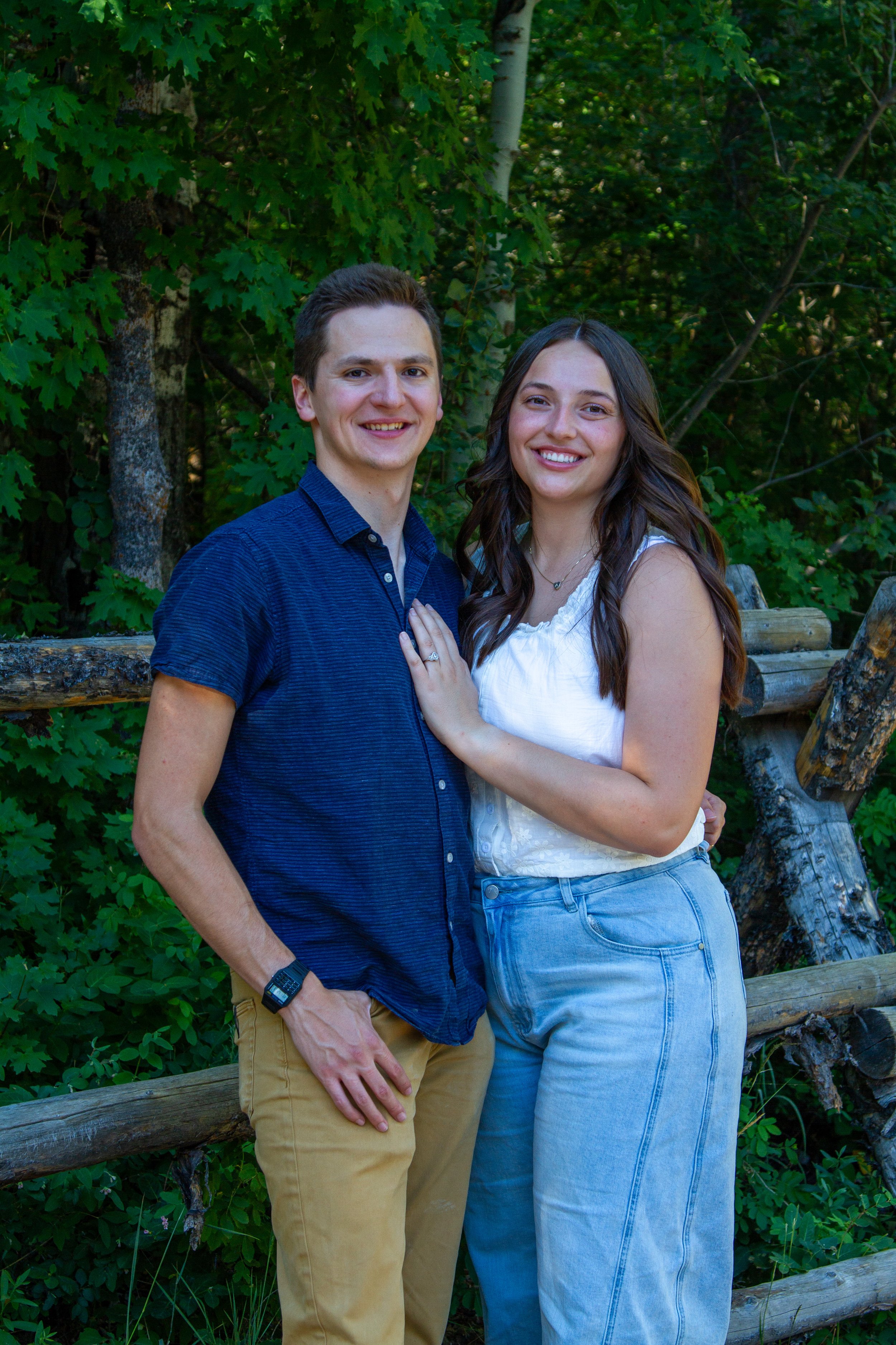 A young man and woman smiling and standing close together outdoors, with green trees and wooden logs in the background.