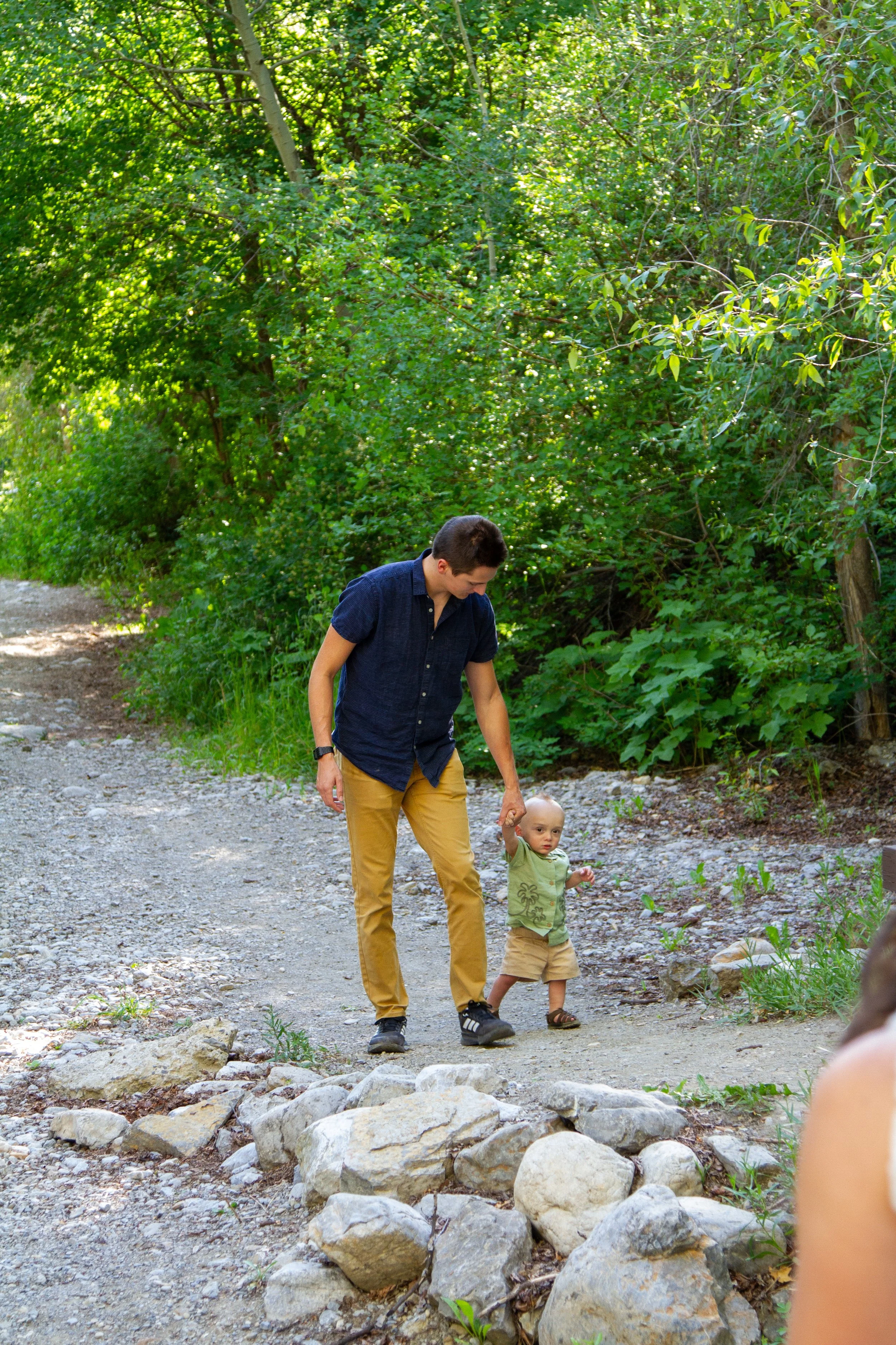 A man and a young child walking on a rocky dirt trail surrounded by lush green trees and bushes in a forest.