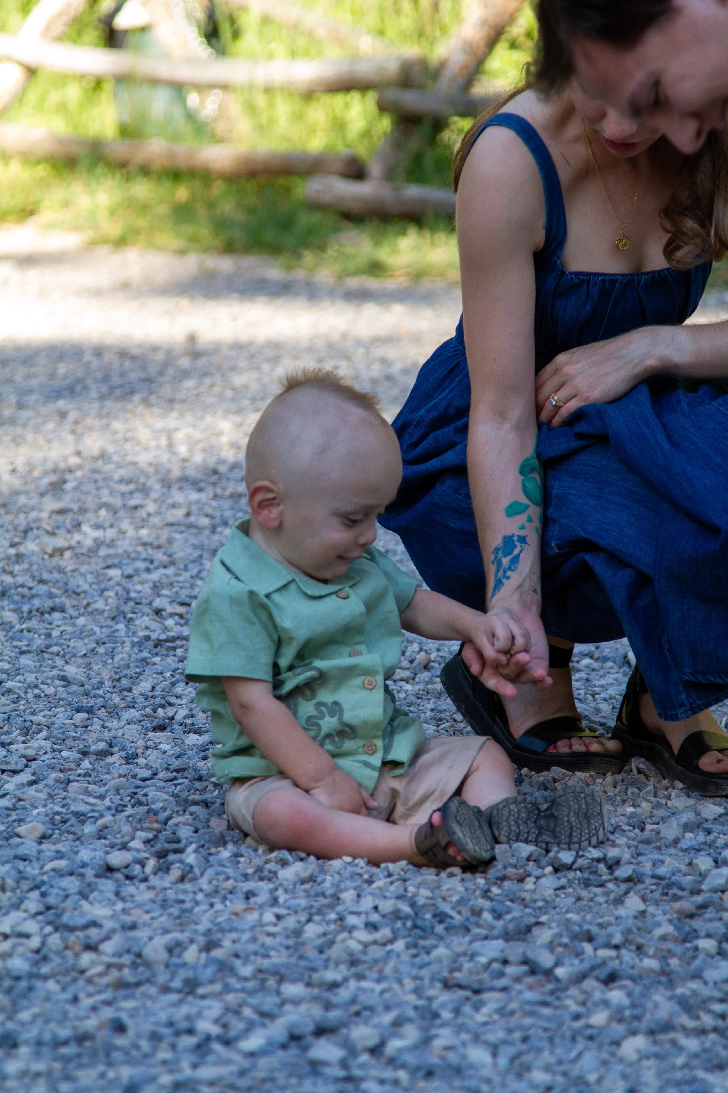 A woman and two children sitting on gravel outdoors, holding hands, with a wooden fence and greenery in the background.