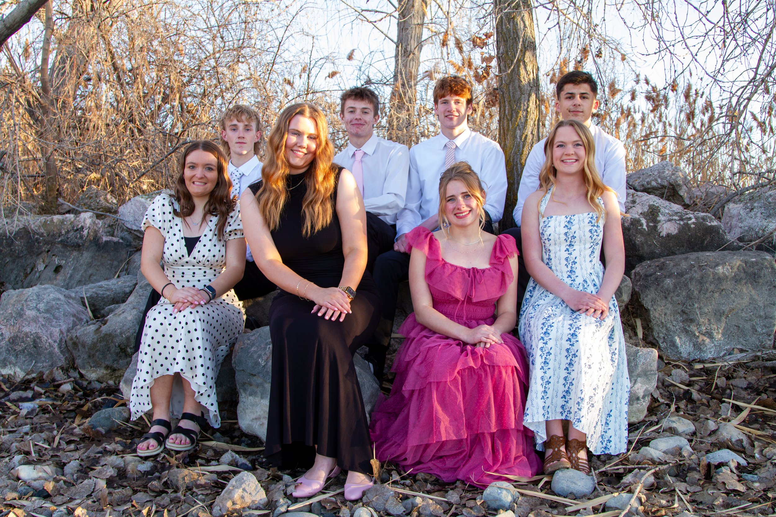 Group of eight young adults, five women and three men, sitting on rocks outdoors in fall, dressed in semi-formal attire, smiling at the camera.