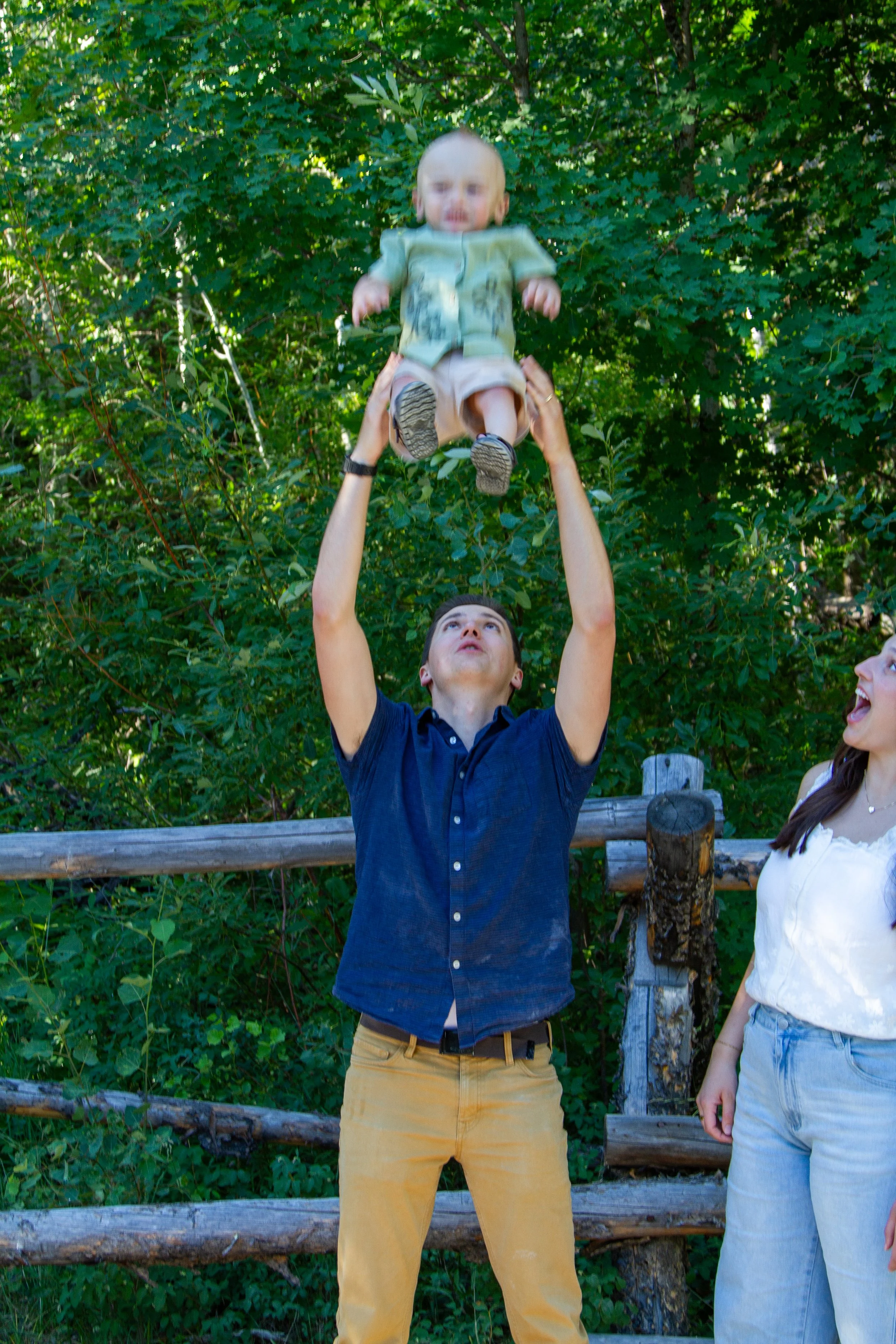A man is lifting a young child into the air outdoors among green trees. The man is wearing a navy blue shirt and khaki pants, and the child is wearing a green shirt and beige shorts. There is a woman standing nearby, partially visible on the right.