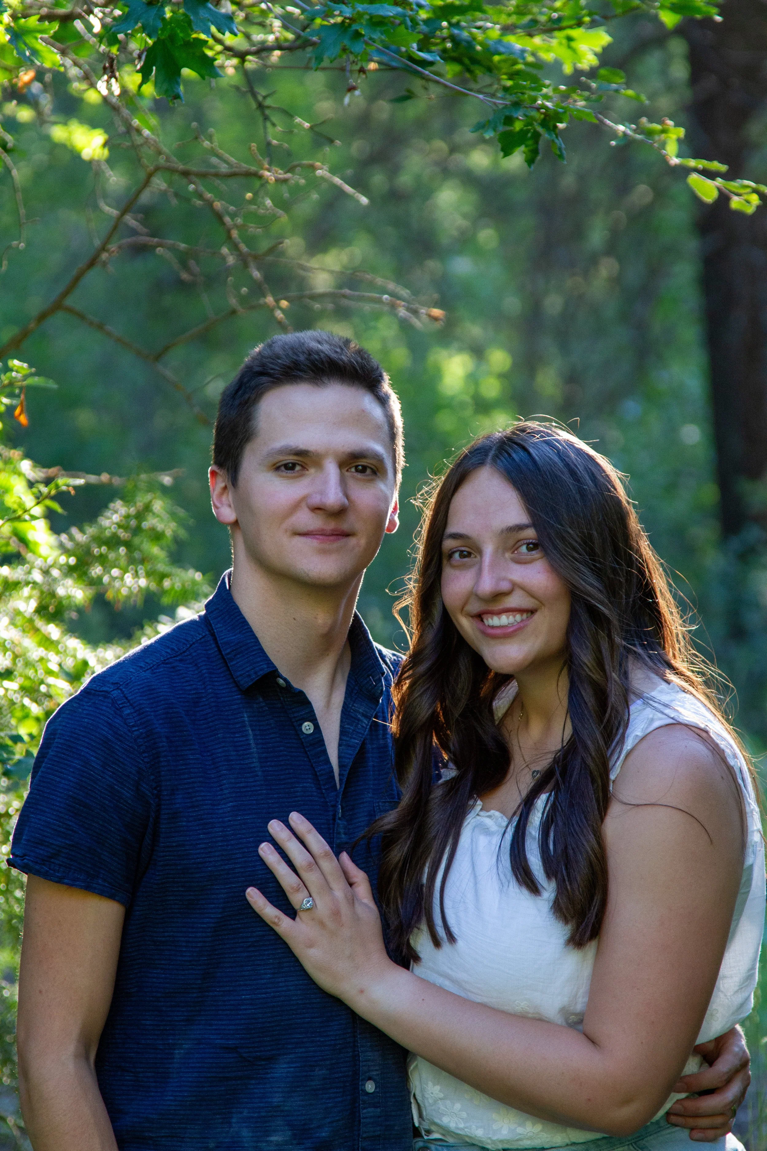 A young man and woman standing close together outdoors in a green, leafy park or garden, smiling at the camera. The man has short dark hair and is wearing a dark blue, short-sleeved, button-up shirt. The woman has long wavy dark hair and is wearing a