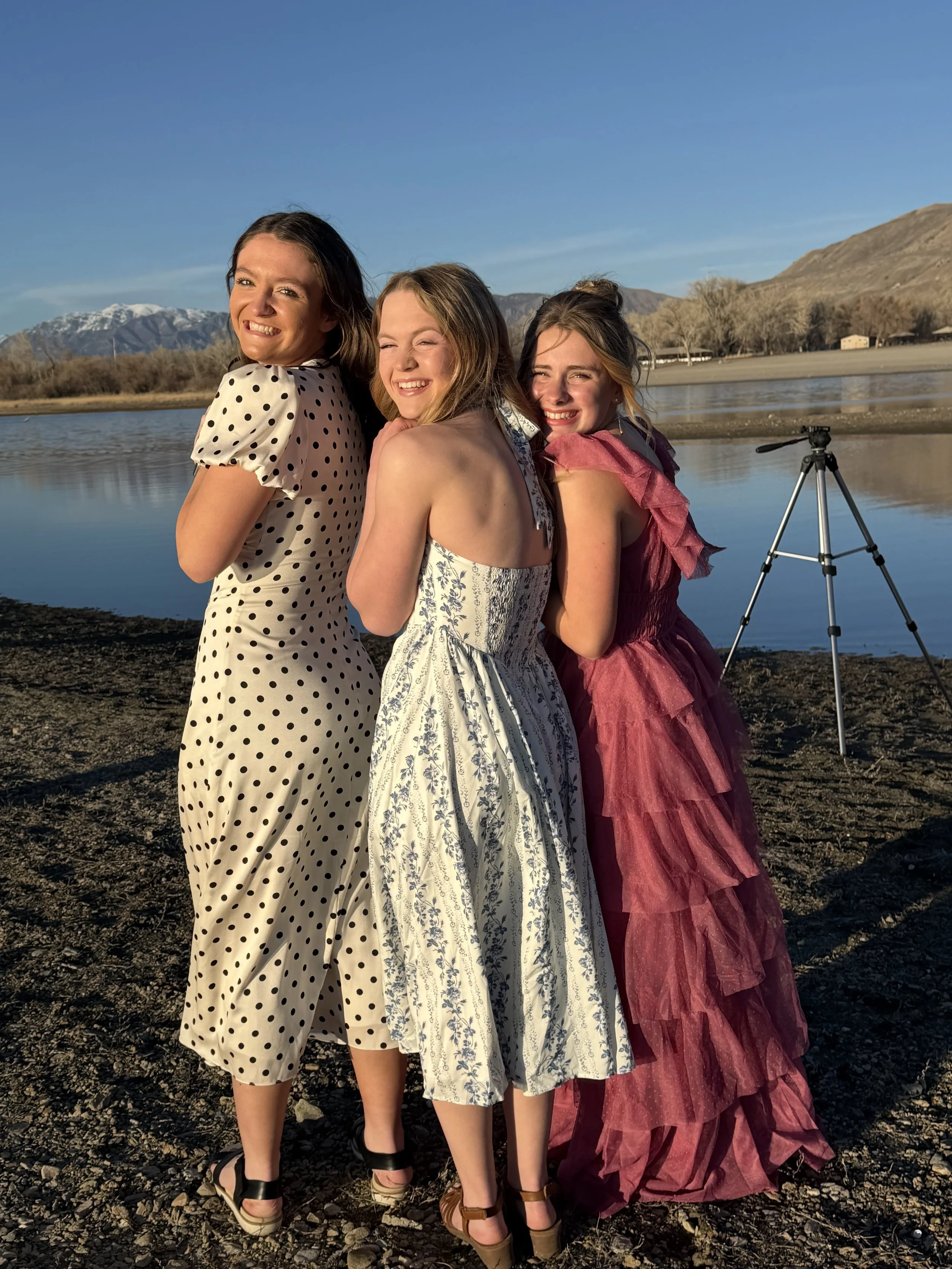 Three young women wearing elegant dresses standing by a lake with mountains in the background, smiling and posing for a photo with a tripod nearby, in sunlight.