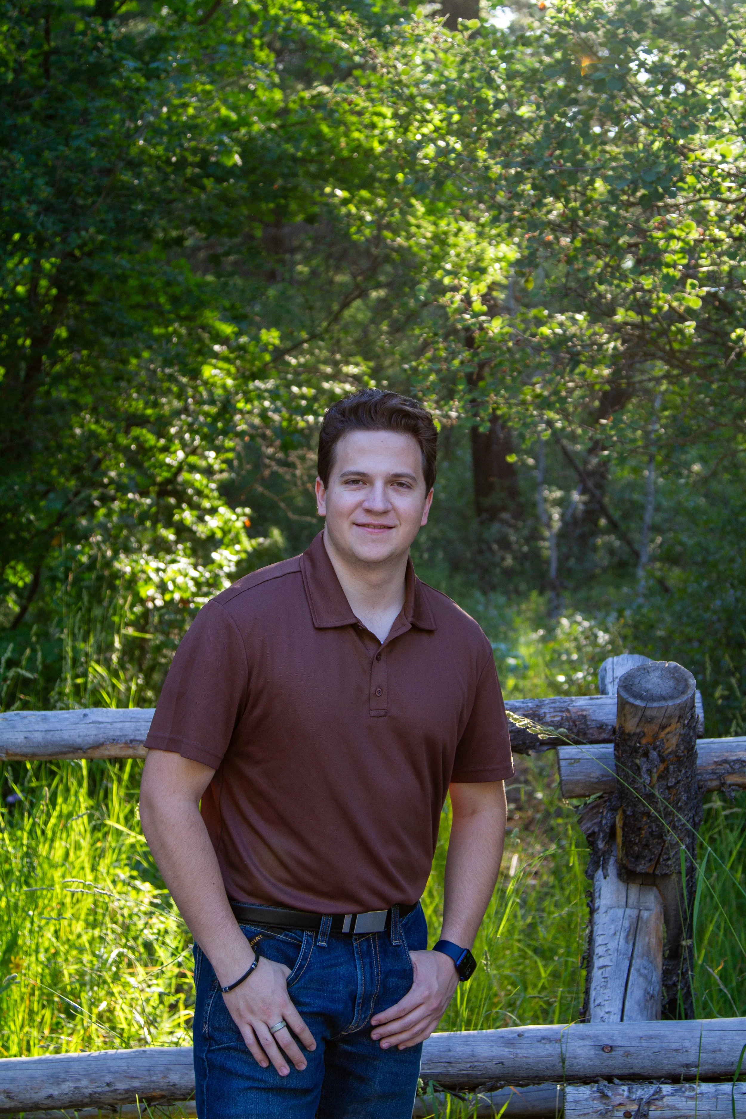 A young man with dark hair, wearing a brown polo shirt and jeans, standing outdoors in front of a wooden fence with green trees and grass in the background, smiling at the camera.