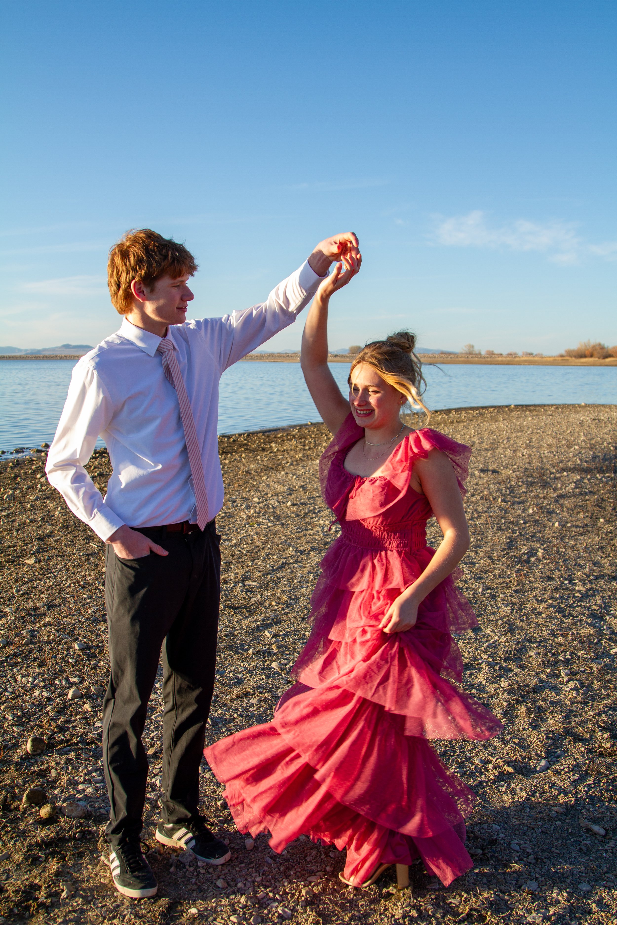 A young man in formal attire and a young woman in a red dress dance on a rocky riverside under a clear blue sky as the sun sets.