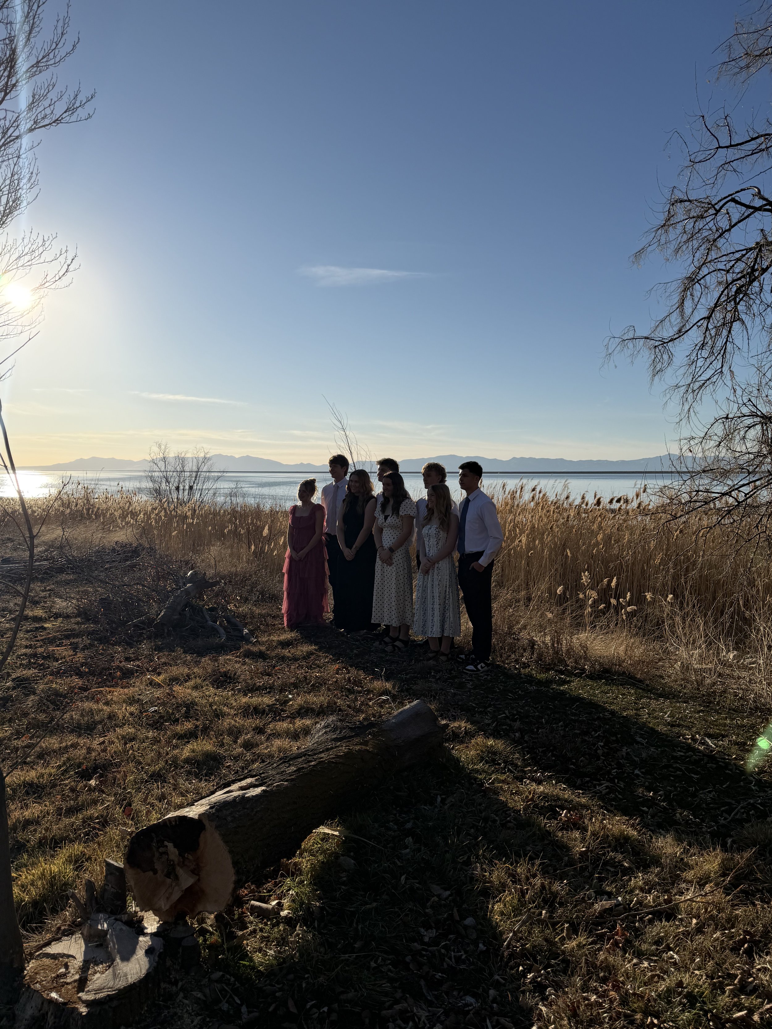 Group of people standing outdoors near a body of water with mountains in the background, during sunset or sunrise.