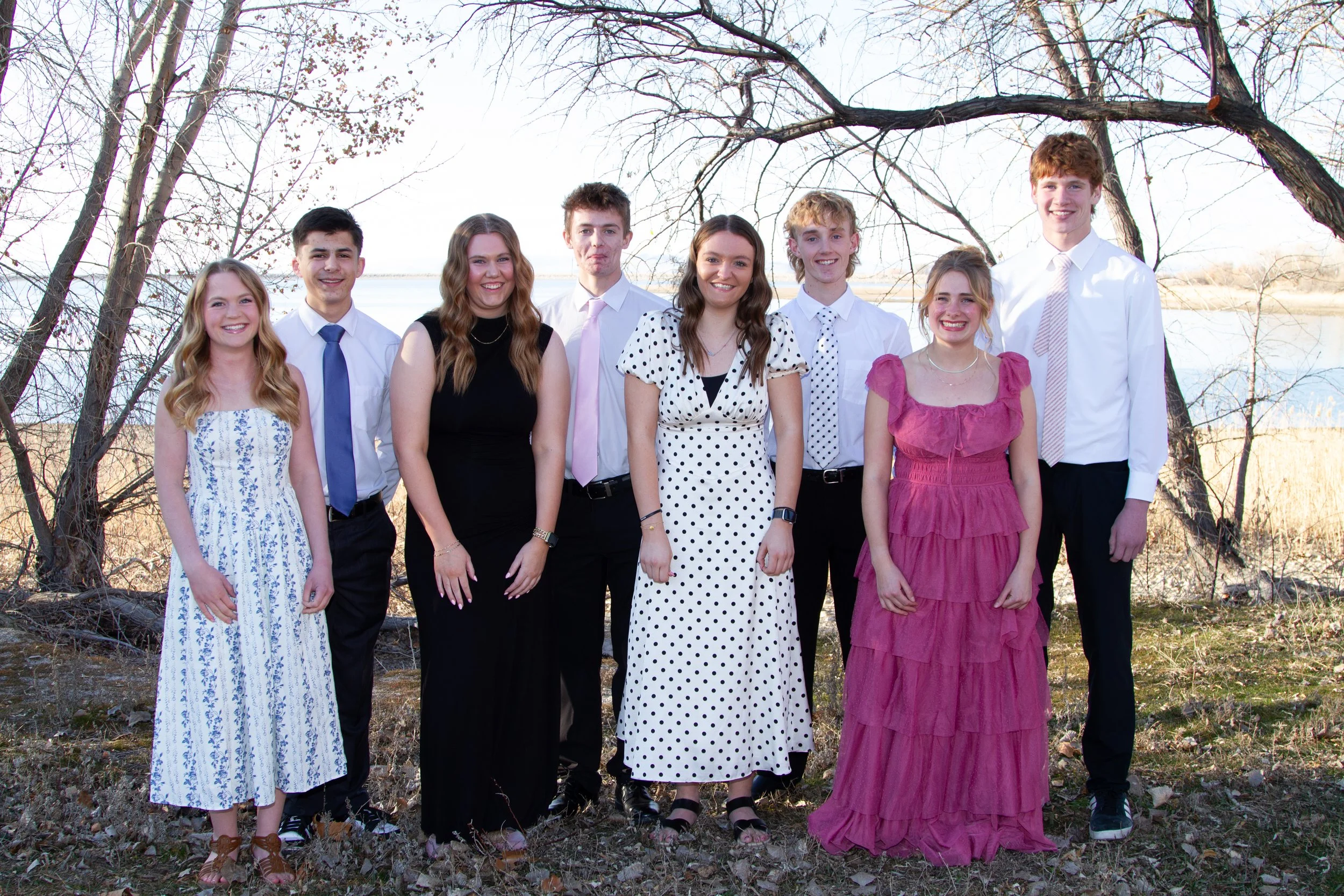 A group of nine young people standing outdoors next to a body of water, dressed in semi-formal attire, under leafless trees, during daytime.