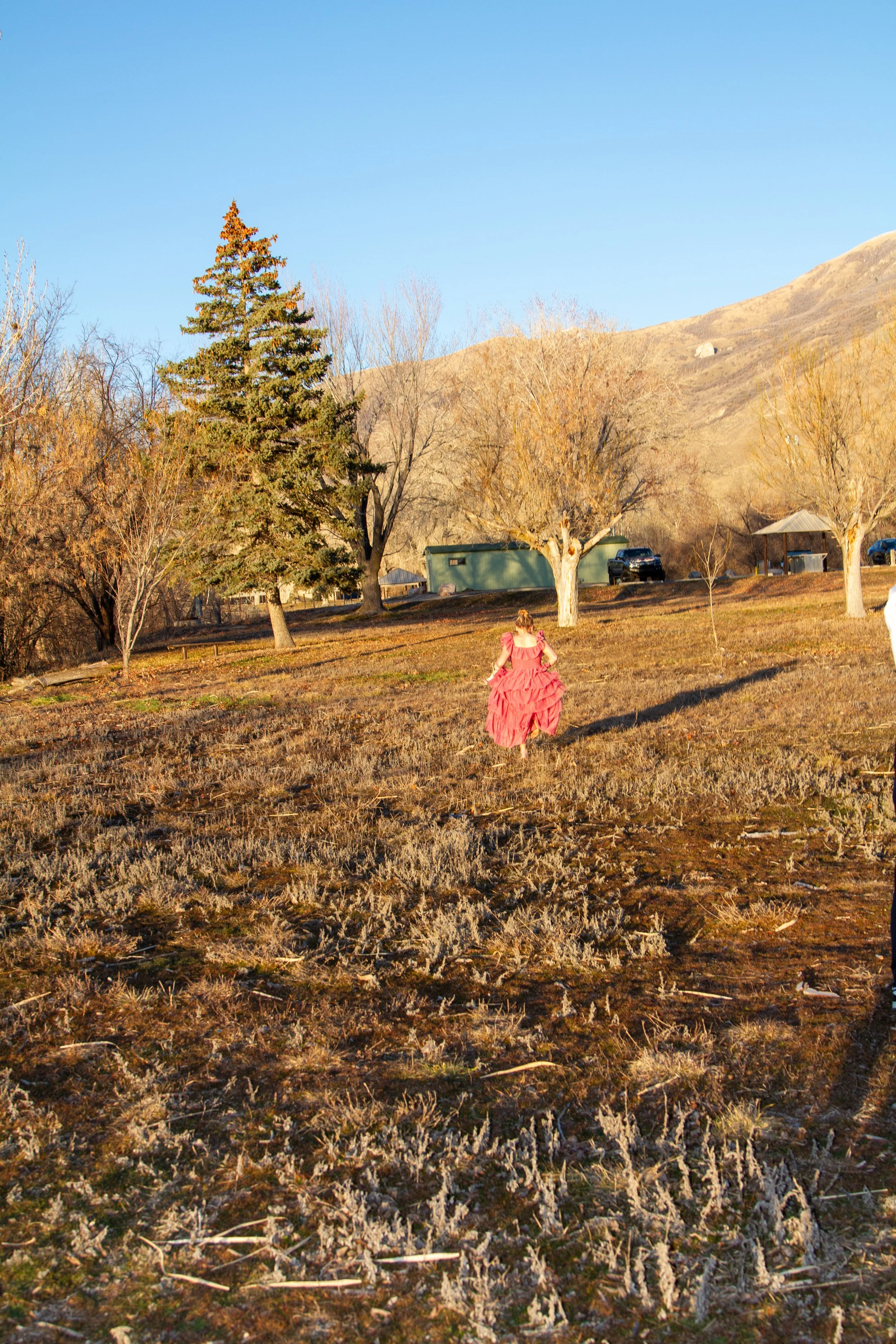 A girl in a pink dress running across a dry, grassy field with trees and hills in the background, under a clear blue sky.