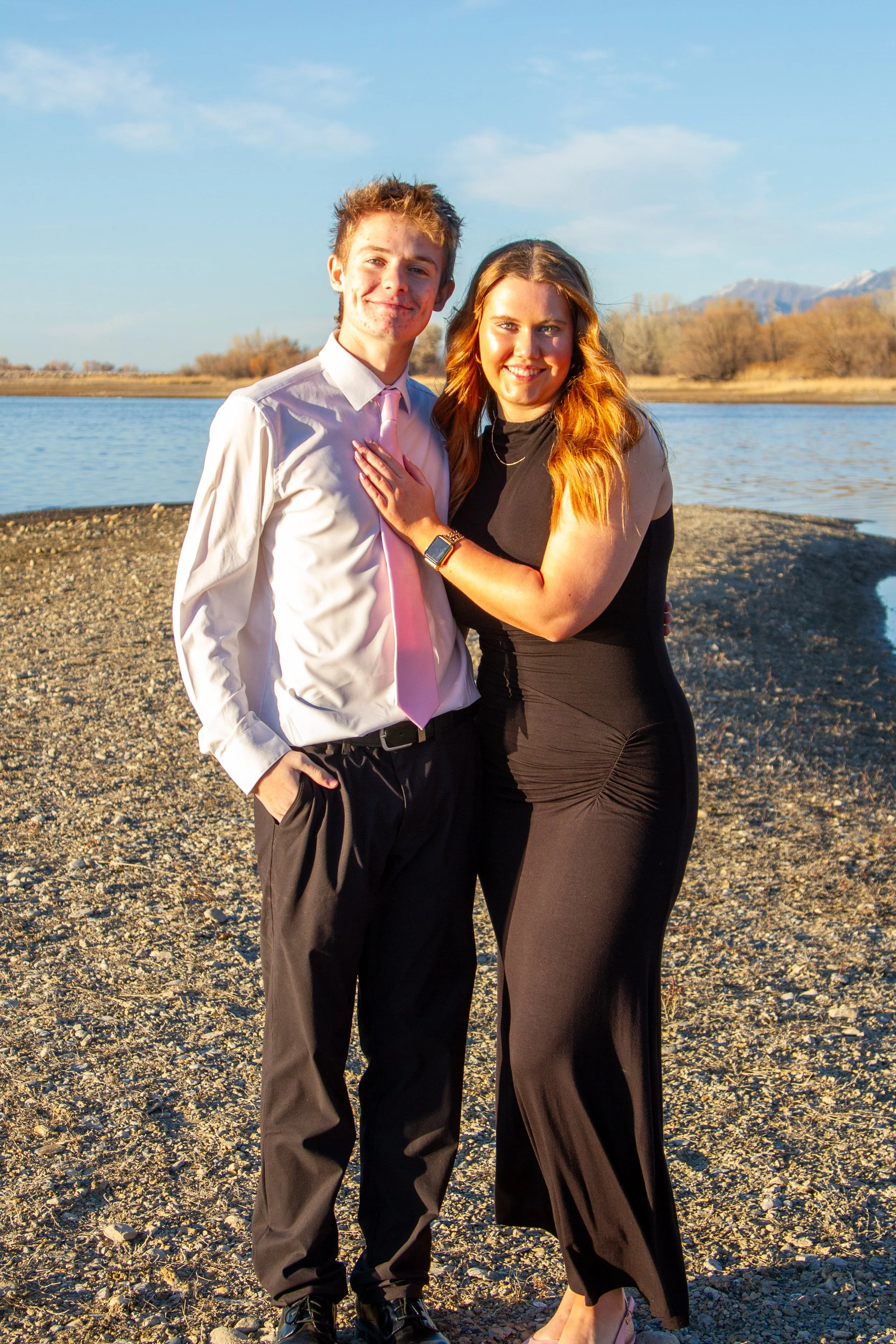 A young couple stands by a body of water outdoors during sunset, smiling at the camera. The man wears a white shirt with a pink tie and dark pants, while the woman wears a black dress.
