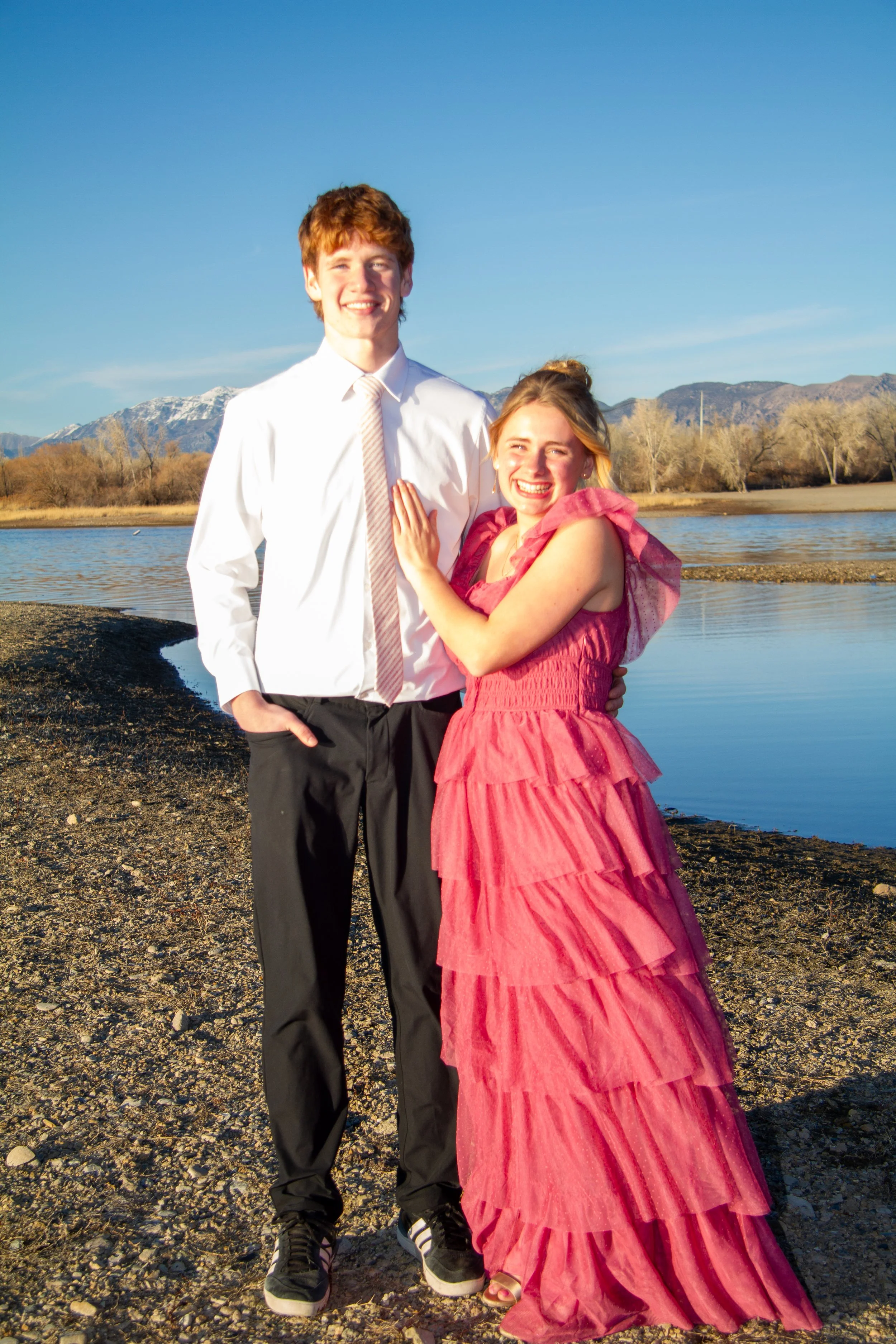 A young man and woman standing together on a rocky riverbank, smiling at the camera. The man is dressed in a white shirt and black pants, the woman in a pink ruffled dress. Mountains are visible in the background under a clear blue sky.
