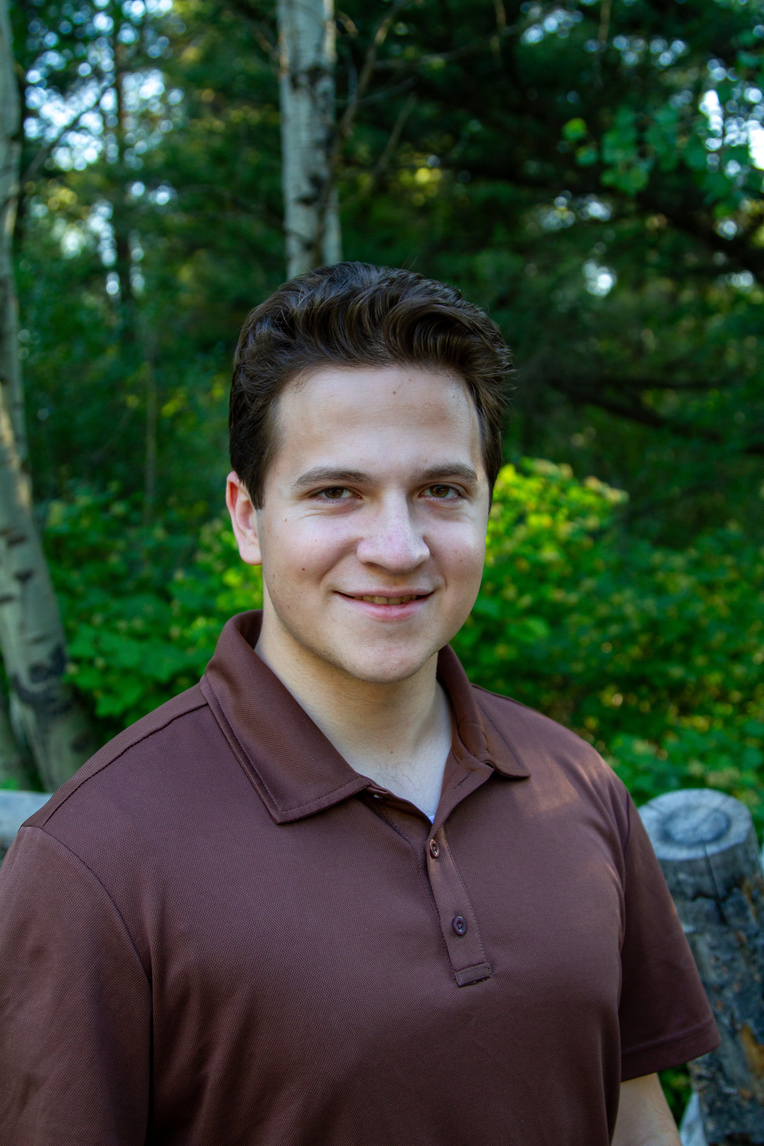 Close-up of a young man with dark hair, smiling, wearing a brown collared shirt, standing outdoors with green trees and foliage in the background.