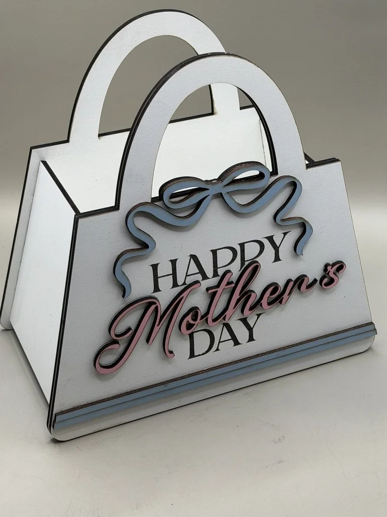 Decorative white shopping bag-shaped sign with the message "Happy Mother's Day" in black and pink lettering, adorned with a blue bow.
