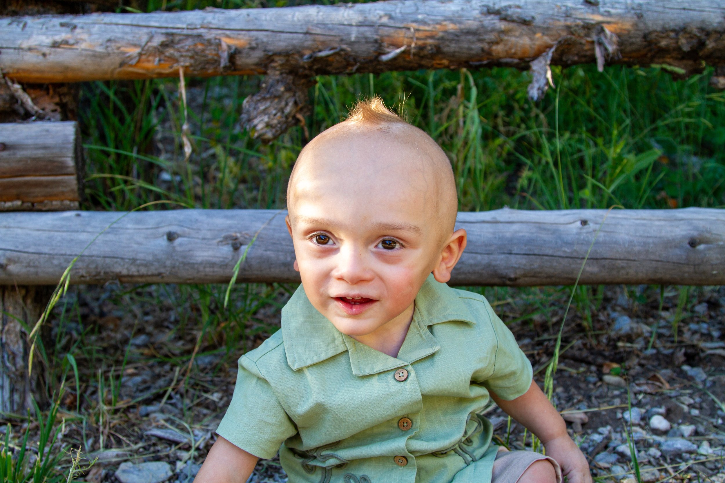 A young boy with a shaved head and light skin, wearing a light green shirt, sitting outdoors on the ground near a wooden fence with green grass and plants behind him.
