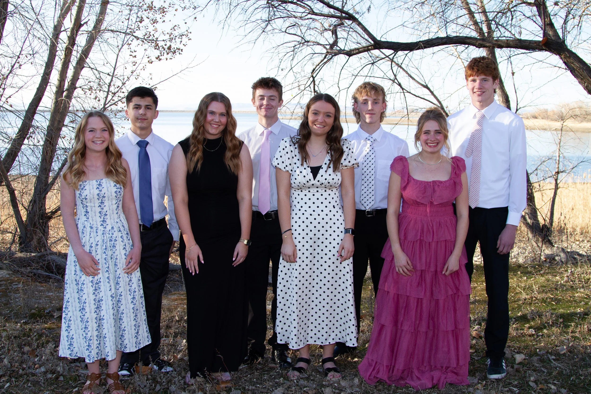 Group of nine young people standing outdoors near a body of water with leafless trees in the background, smiling and dressed in semi-formal attire.