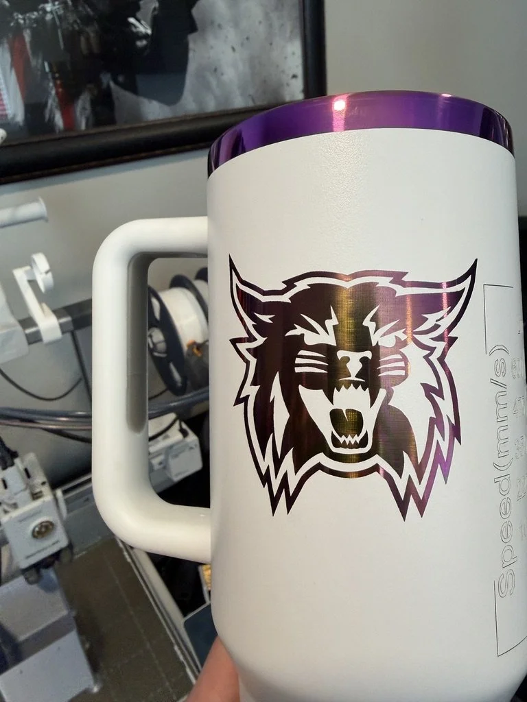 A white ceramic mug with a purple interior, featuring a large black lion head logo on the front, held by a person in a workshop or industrial setting.
