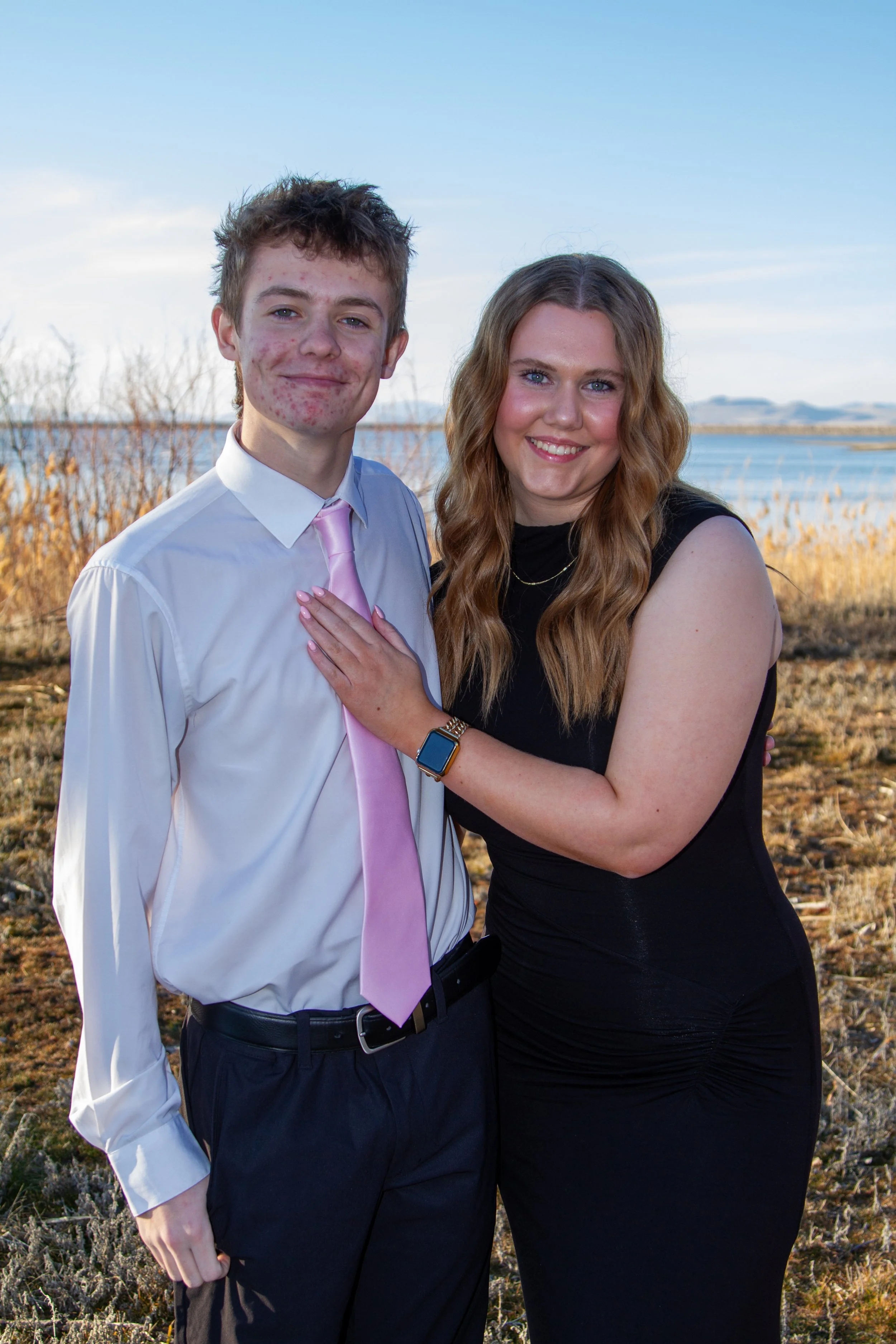 A young man and a woman standing outdoors near a body of water, smiling at the camera during daytime.