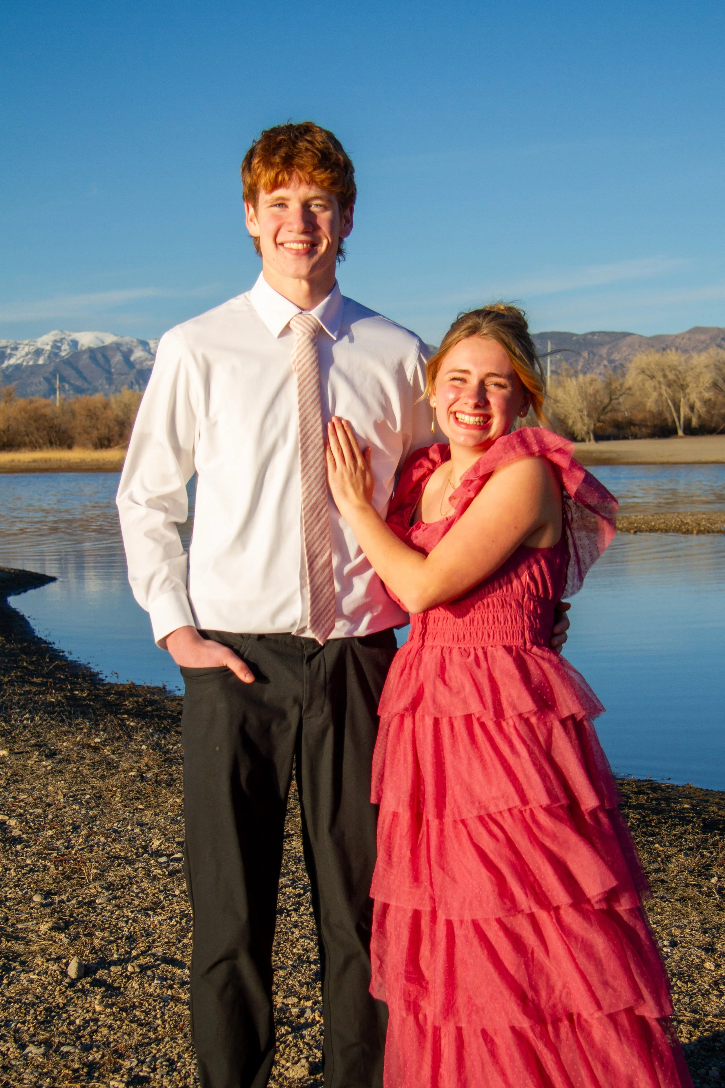 A young man and woman standing together outdoors near a river, smiling. The man is wearing a white dress shirt and black trousers, and the woman is dressed in a ruffled red gown. Snow-capped mountains are visible in the background under a clear blue 