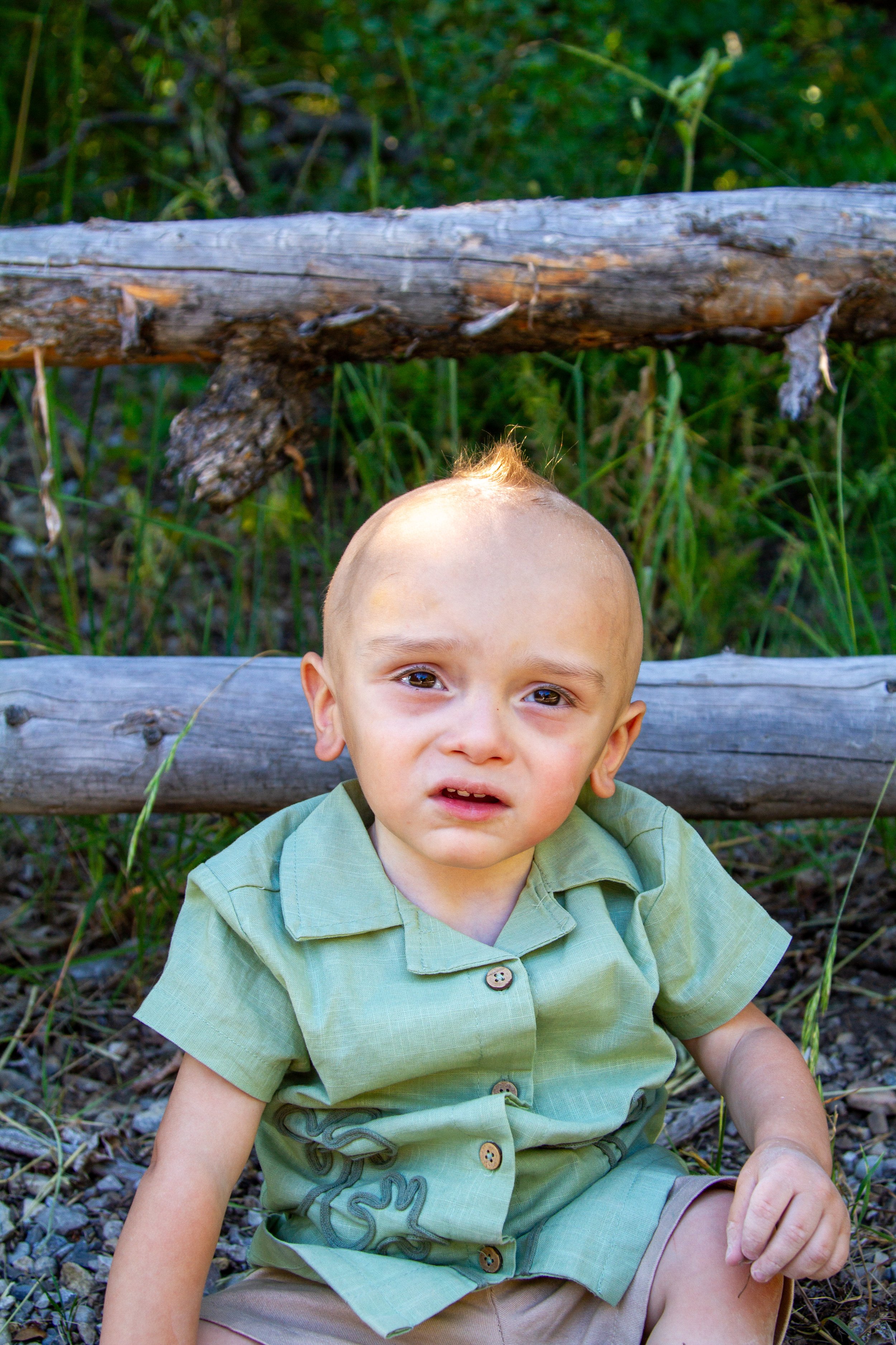 A young boy with light skin, short light brown hair, and brown eyes sitting outdoors in front of a wooden fence with green foliage. He's wearing a light green button-up shirt with rolled-up sleeves and tan shorts. The boy is looking at the camera wit