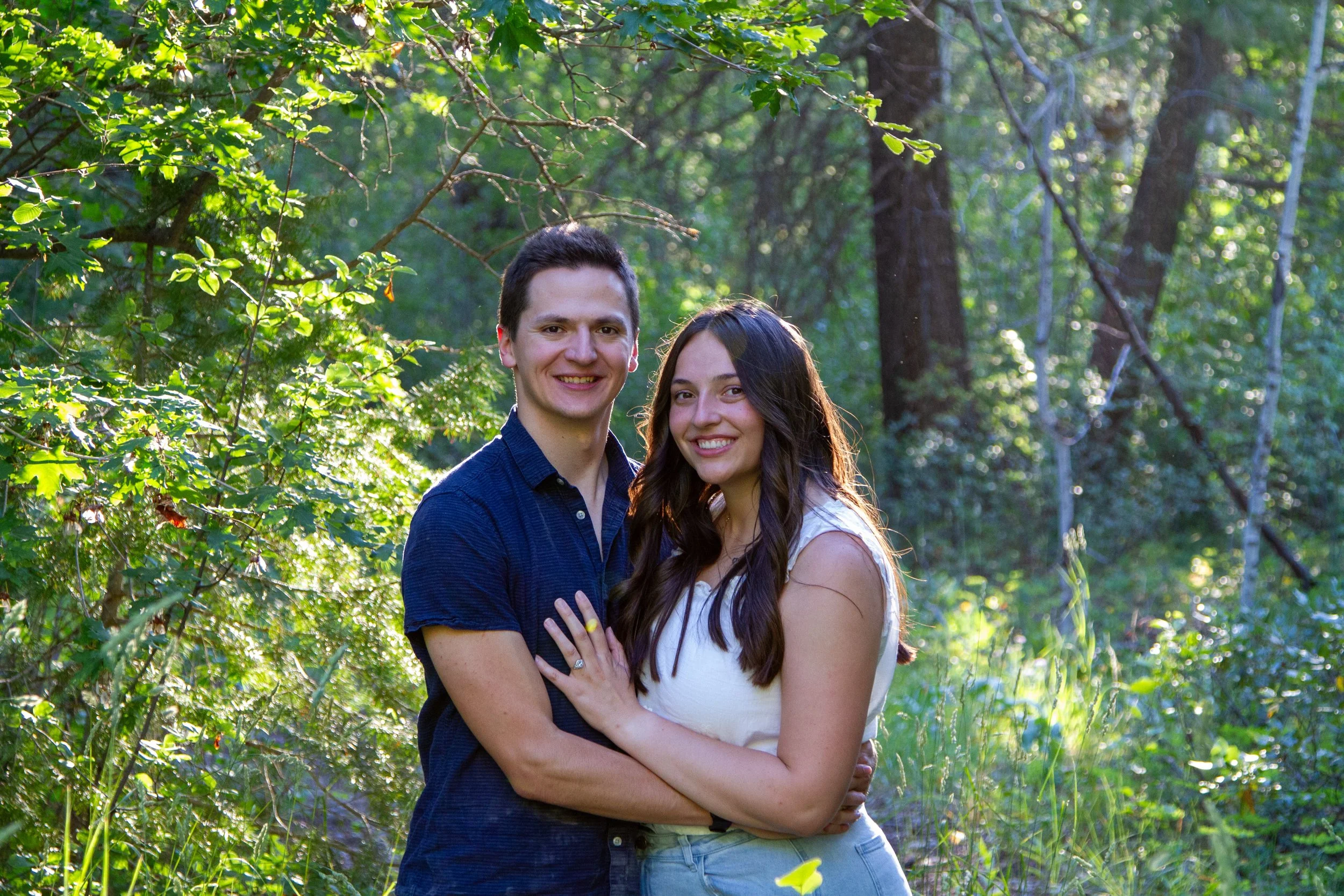 A young couple stands closely together outdoors in a lush green forest, smiling at the camera. The man has dark hair and wears a blue shirt, while the woman has long dark hair and wears a white top. They are embracing each other amidst trees and foli