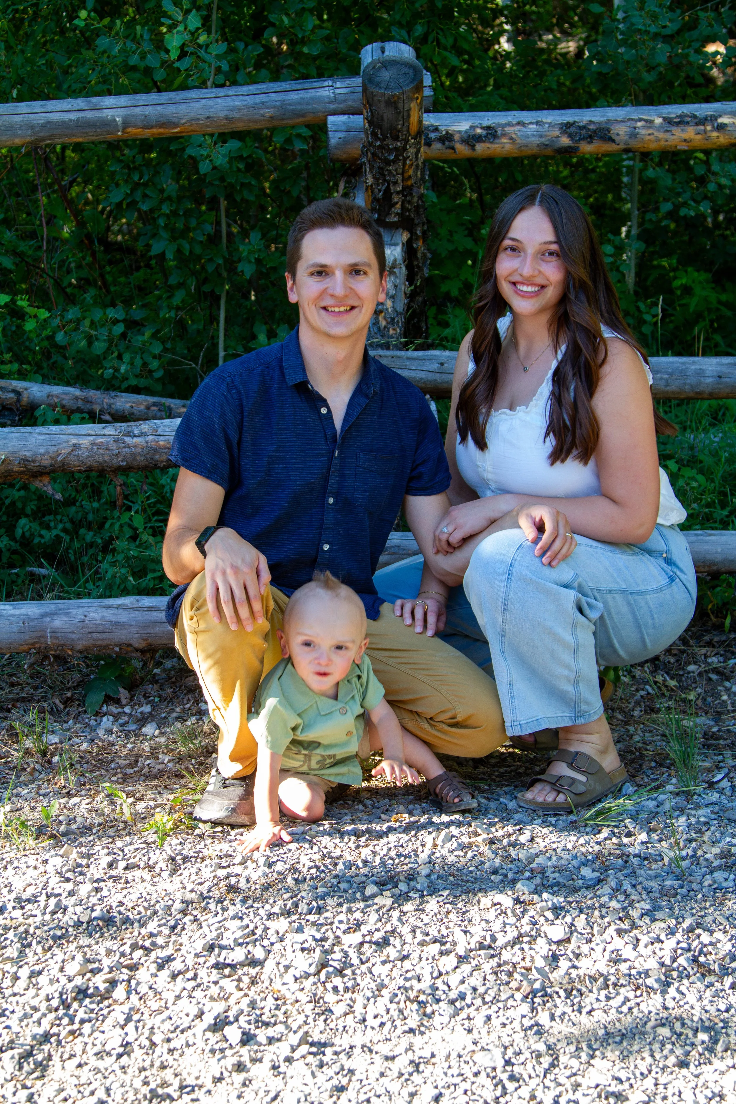 A young man and woman sit on a wooden fence with a toddler outdoors, surrounded by green foliage and gravel ground, smiling at the camera.