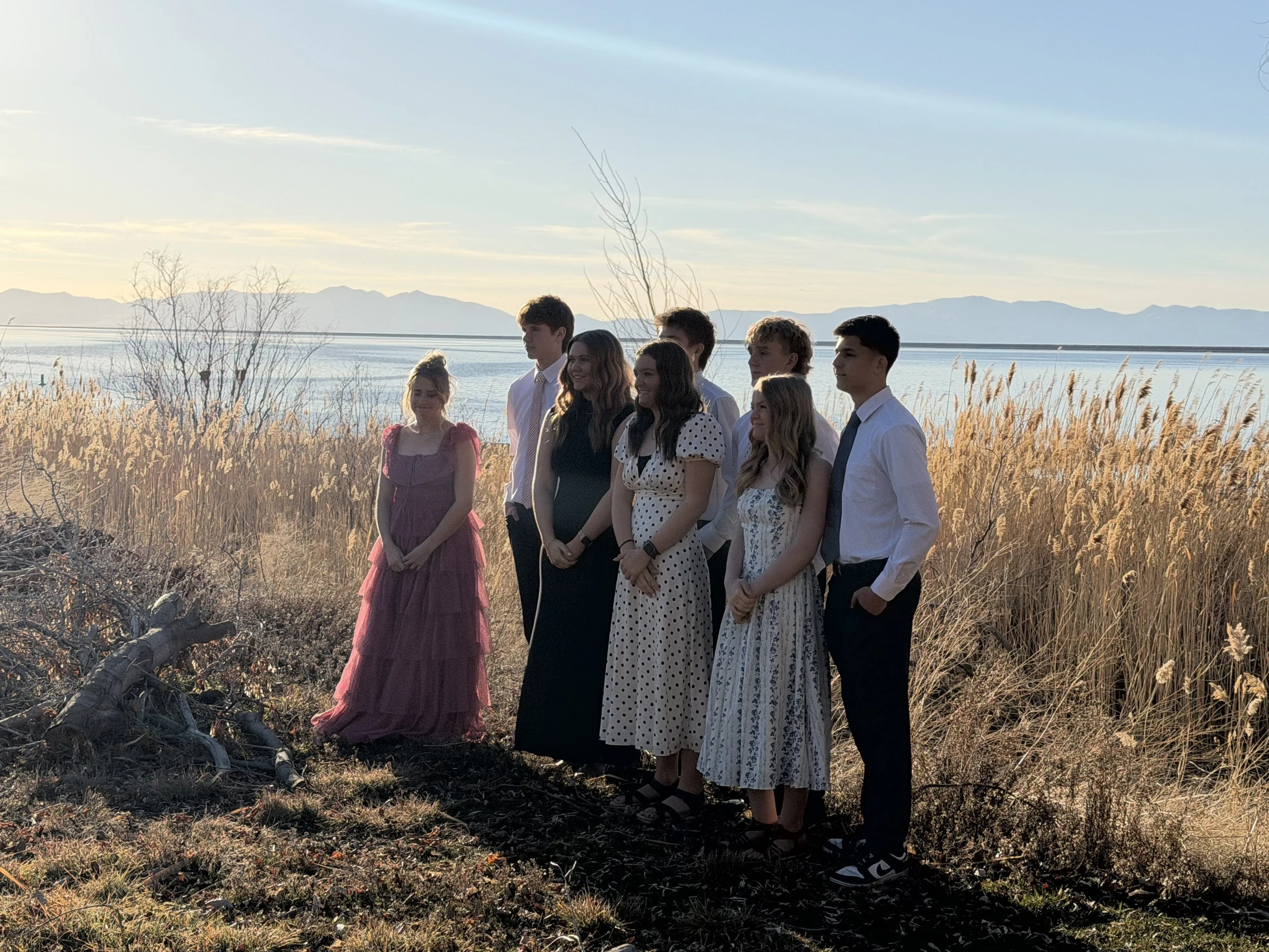 Group of young people standing outdoors near a lake or river with mountains in the distance, wearing semi-formal attire during sunset.