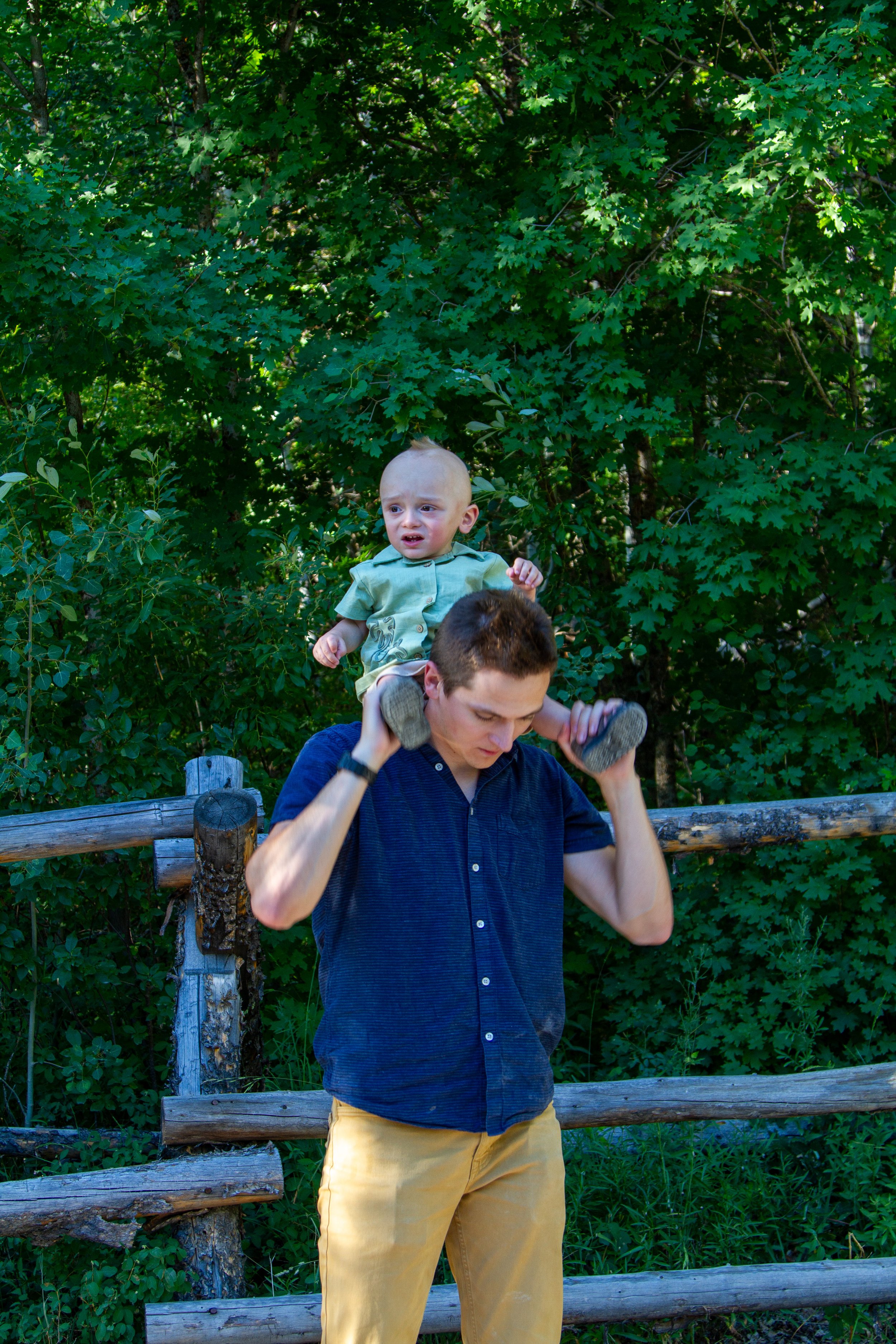 A young man with short brown hair in a navy blue shirt and yellow pants is holding a small child on his shoulders outdoors. The child has light skin and is wearing a light green shirt. They are in front of a wooden fence with lush green trees and fol