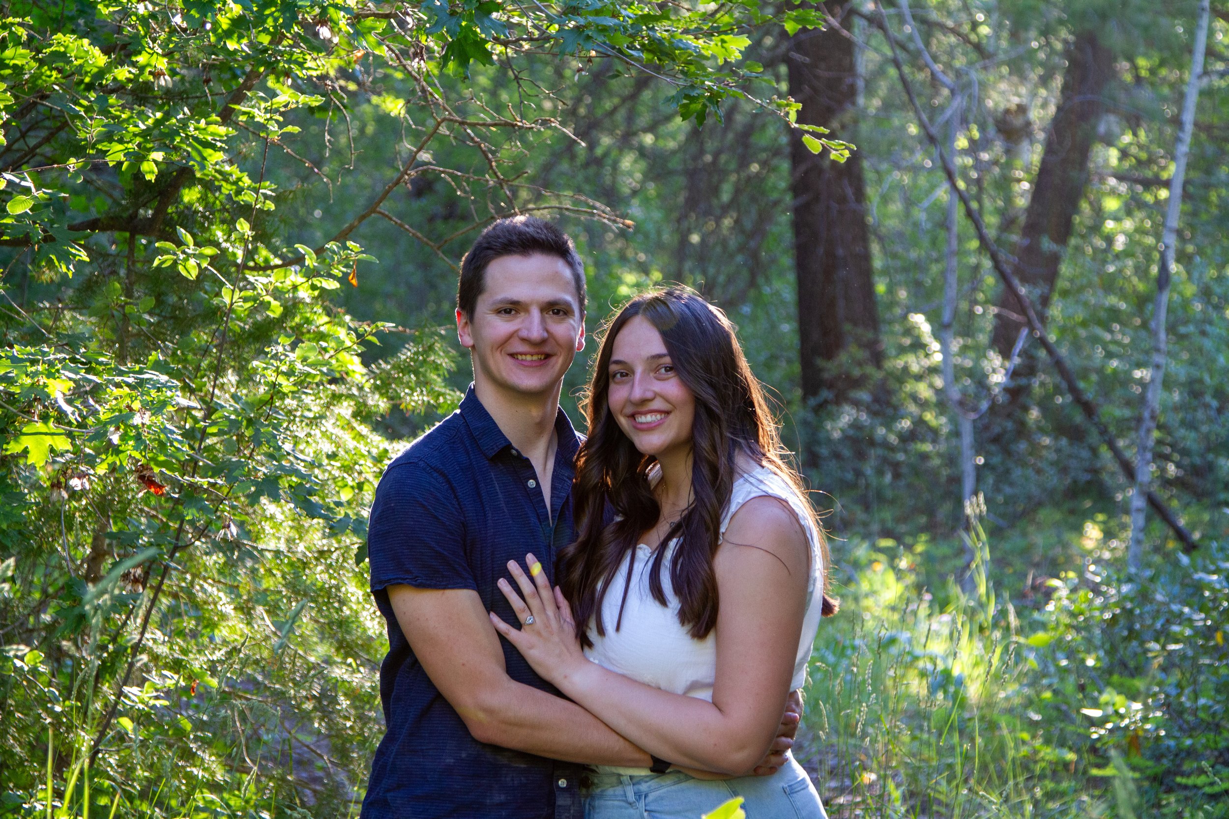 A smiling young man and woman embracing outdoors in a green, wooded area with sunlight filtering through the trees.