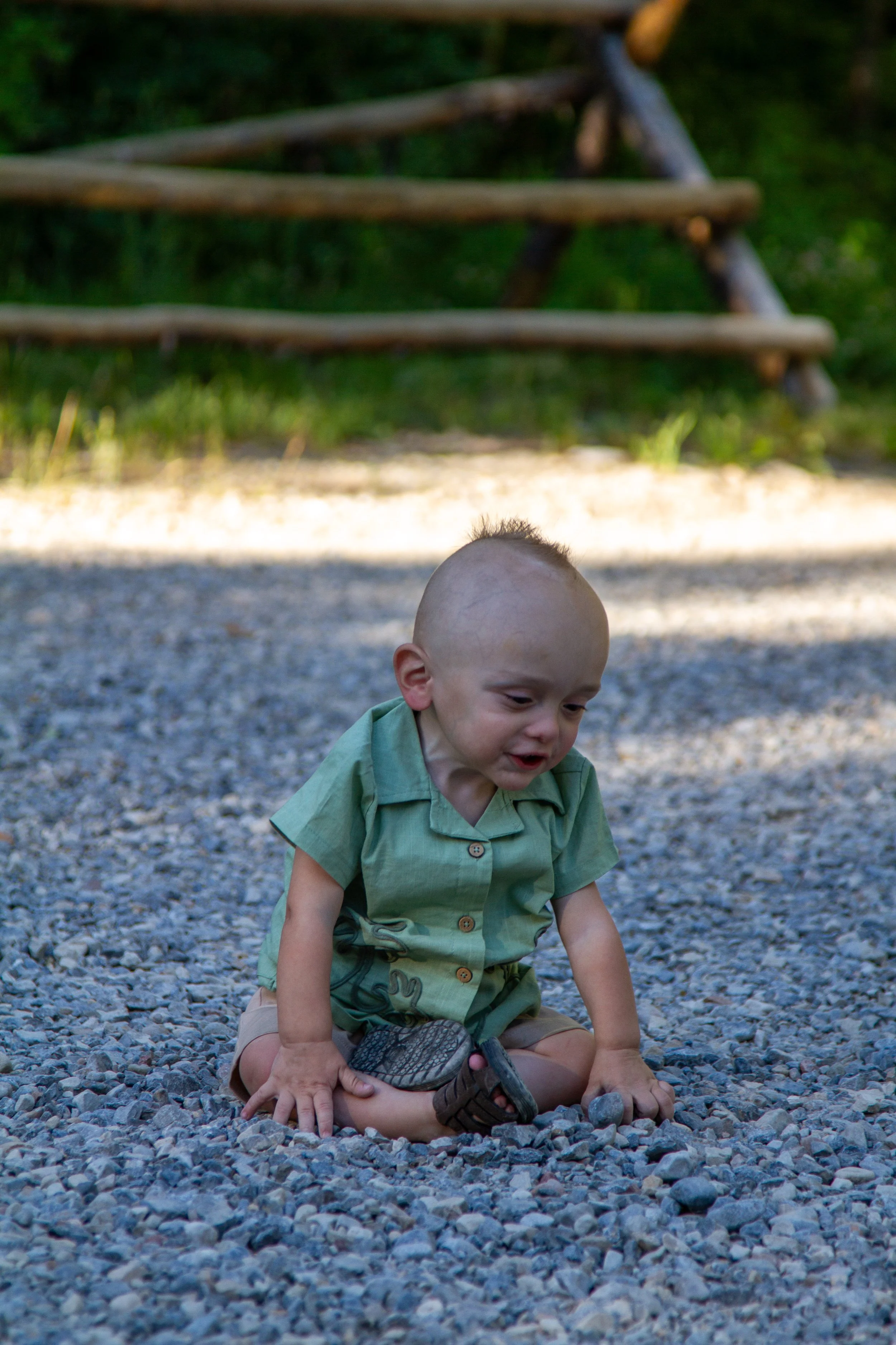A young boy with a shaved head wearing a green shirt and brown shorts, sitting on a gravel surface outdoors with a wooden structure and green foliage in the background.