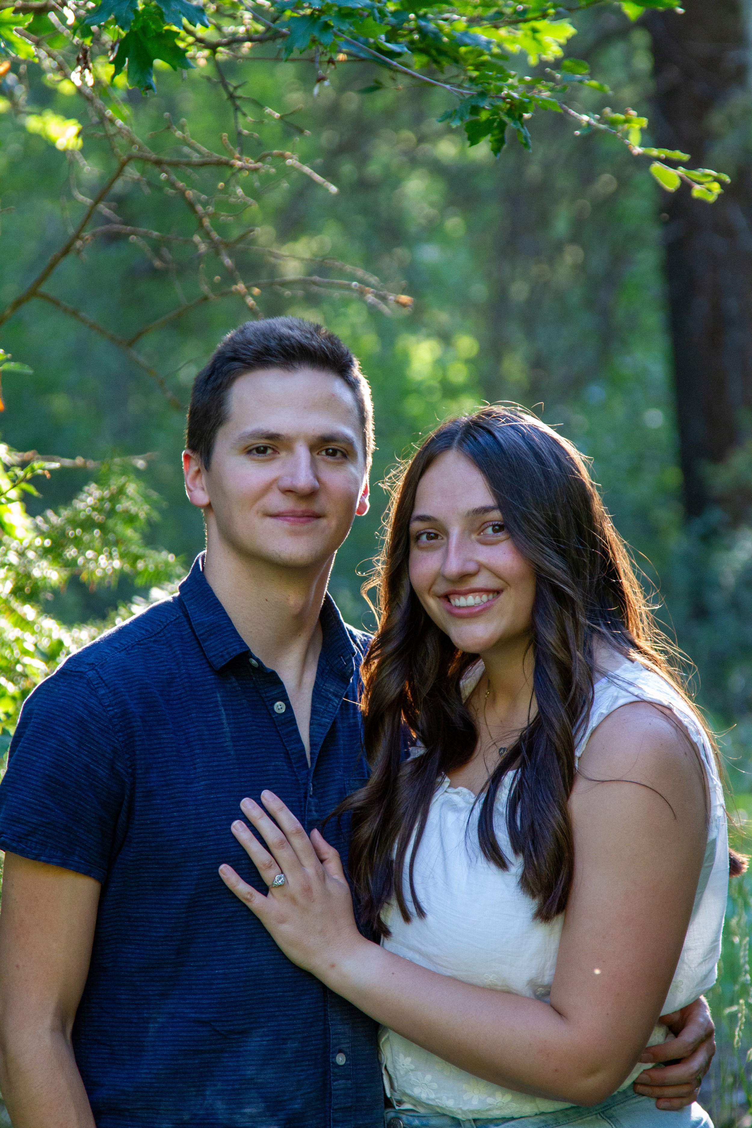 A young man and woman standing close together outdoors in a green, sunlit park with trees and foliage in the background. The woman is smiling and has her arm around the man's waist, and the man's arm is around the woman's shoulder.