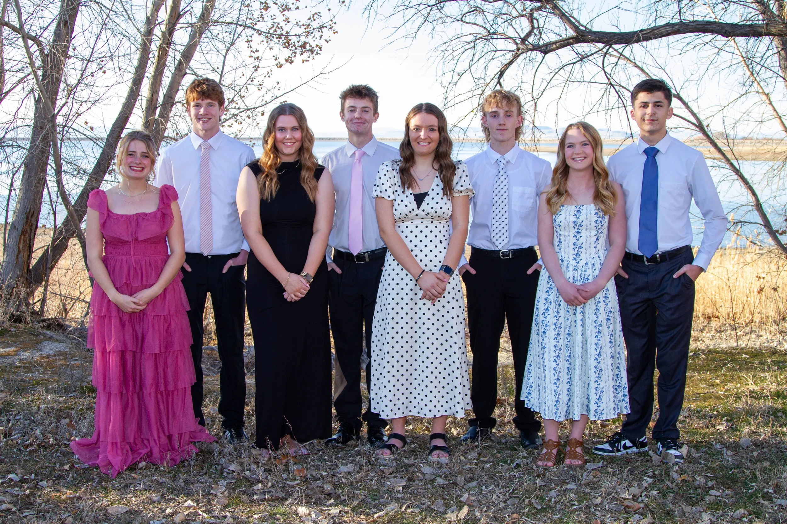 Group of eight young people outdoors near a body of water, dressed in semi-formal attire, standing in front of trees with bare branches.
