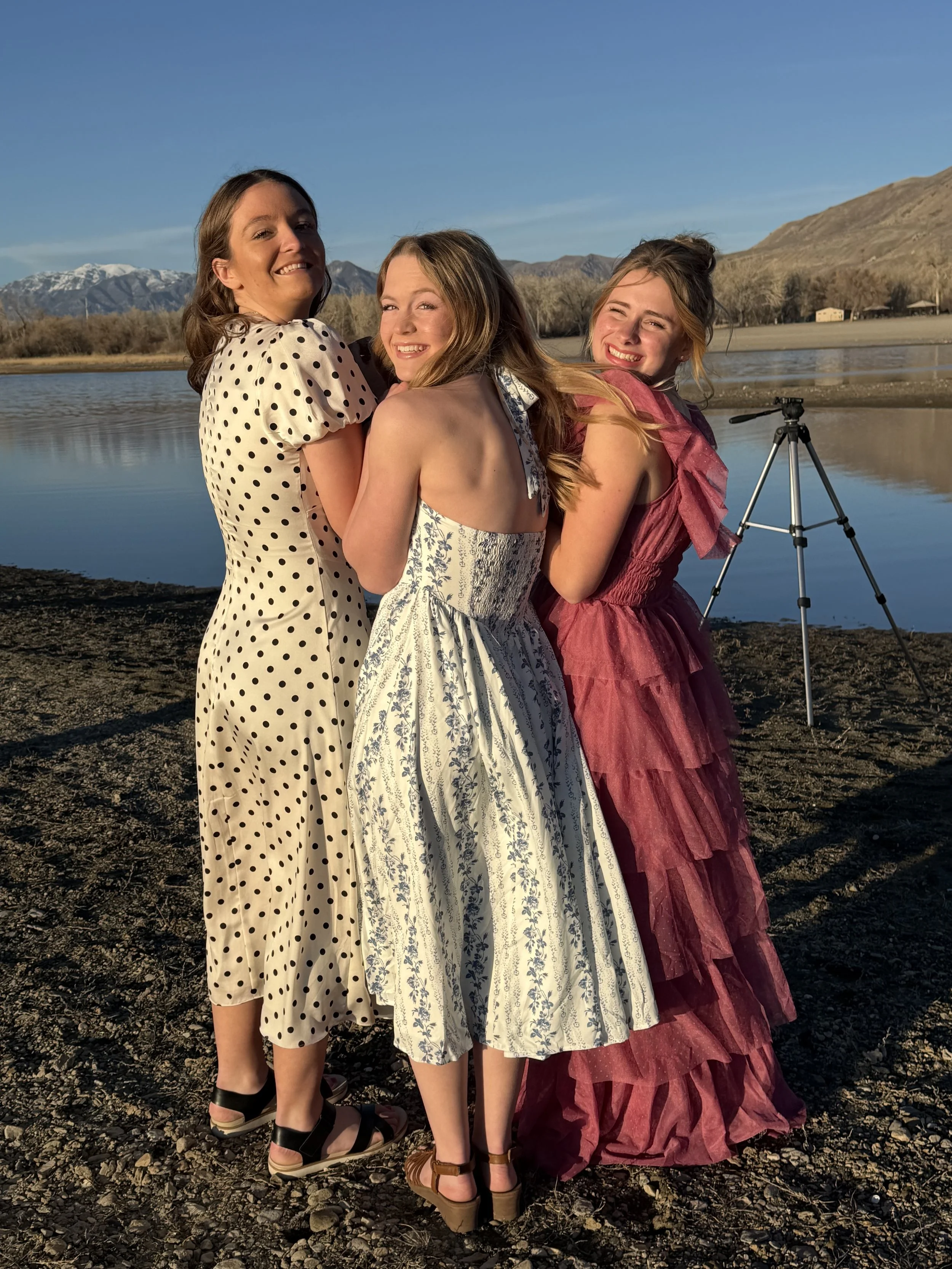 Three young women in dresses hugging and smiling on a rocky shore with a lake, mountains, and a clear sky in the background, and a tripod nearby.