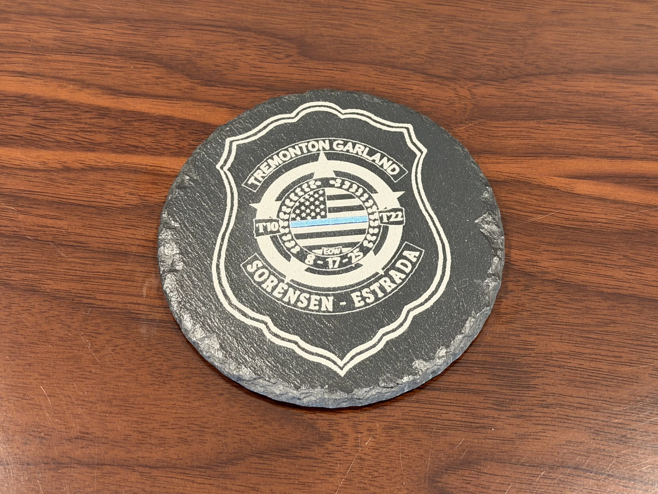 Close-up of a round, black stone coaster with white graphics and text, placed on a wooden table.